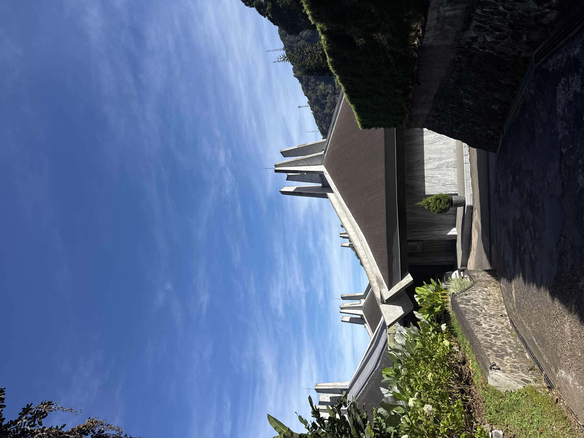 The image shows a building with a modern architectural style against a clear blue sky. The building has a distinctive roofline with several vertical concrete fins extending upwards. The walls appear to be made of concrete. The building is located in an environment with greenery including bushes and trees. A glimpse of what appears to be a hillside or mountain is visible in the background. There are no people visible in the image. There is no information in the image to confirm the location in the city of Unknown, Unknown.