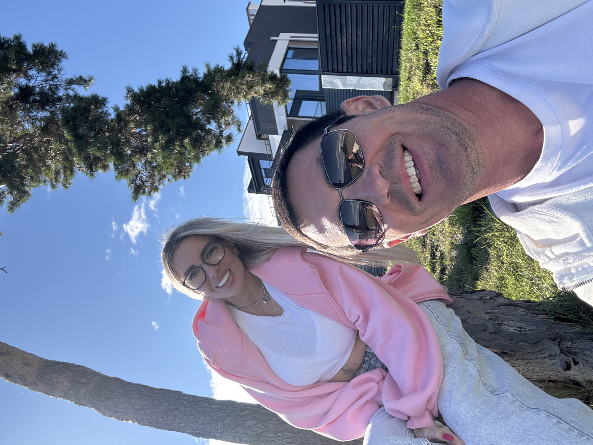 This is a selfie of a man and a woman outdoors, with a modern house and trees in the background. The man is in the foreground, smiling and wearing sunglasses and a white jacket. The woman is behind him, wearing a pink hoodie and glasses, also smiling. The scene appears to be a sunny day. Based on the architectural style and landscaping, this location could be in a residential area, possibly with a view of the sea or a body of water, which is typical of many areas in Primorsky Krai, Russia.