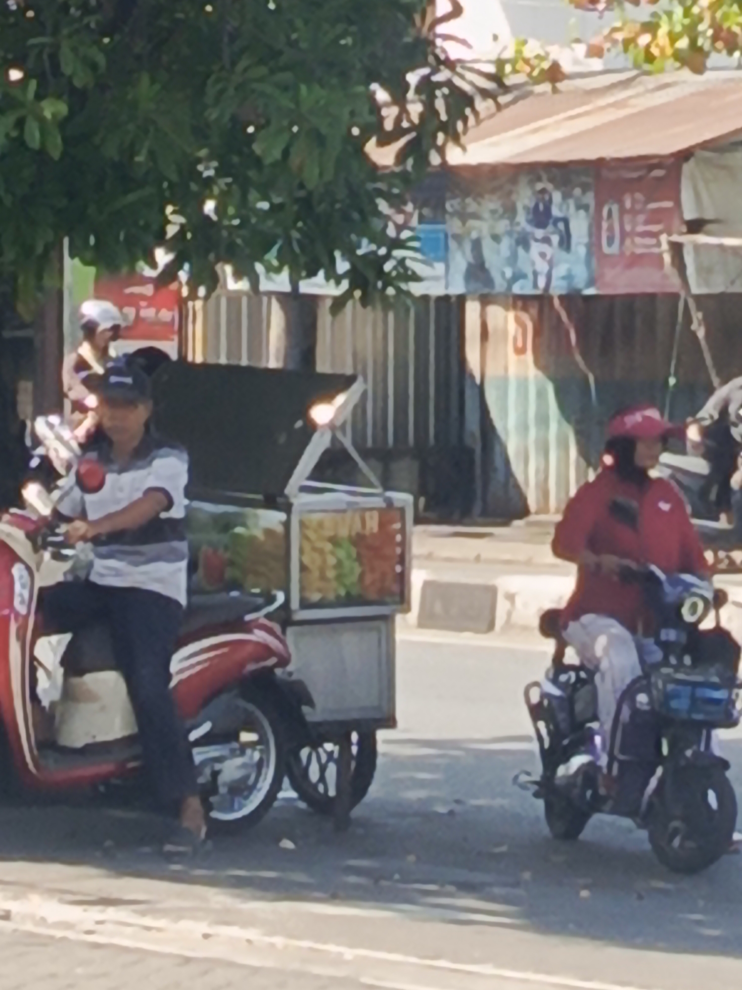 The image captures a street scene in Surabaya, Indonesia. In the foreground, a person wearing a striped shirt and a dark cap is seated on a red and white scooter. To their right, a mobile food cart, possibly selling fruit, is parked. Further to the right, another person wearing a red top and a pink visor is riding a dark-colored scooter. Behind the food cart, a building with a corrugated metal roof is visible, adorned with banners. Trees with green foliage are present in the upper left portion of the image. The scene appears to be during daylight, with shadows indicating the sun's position.