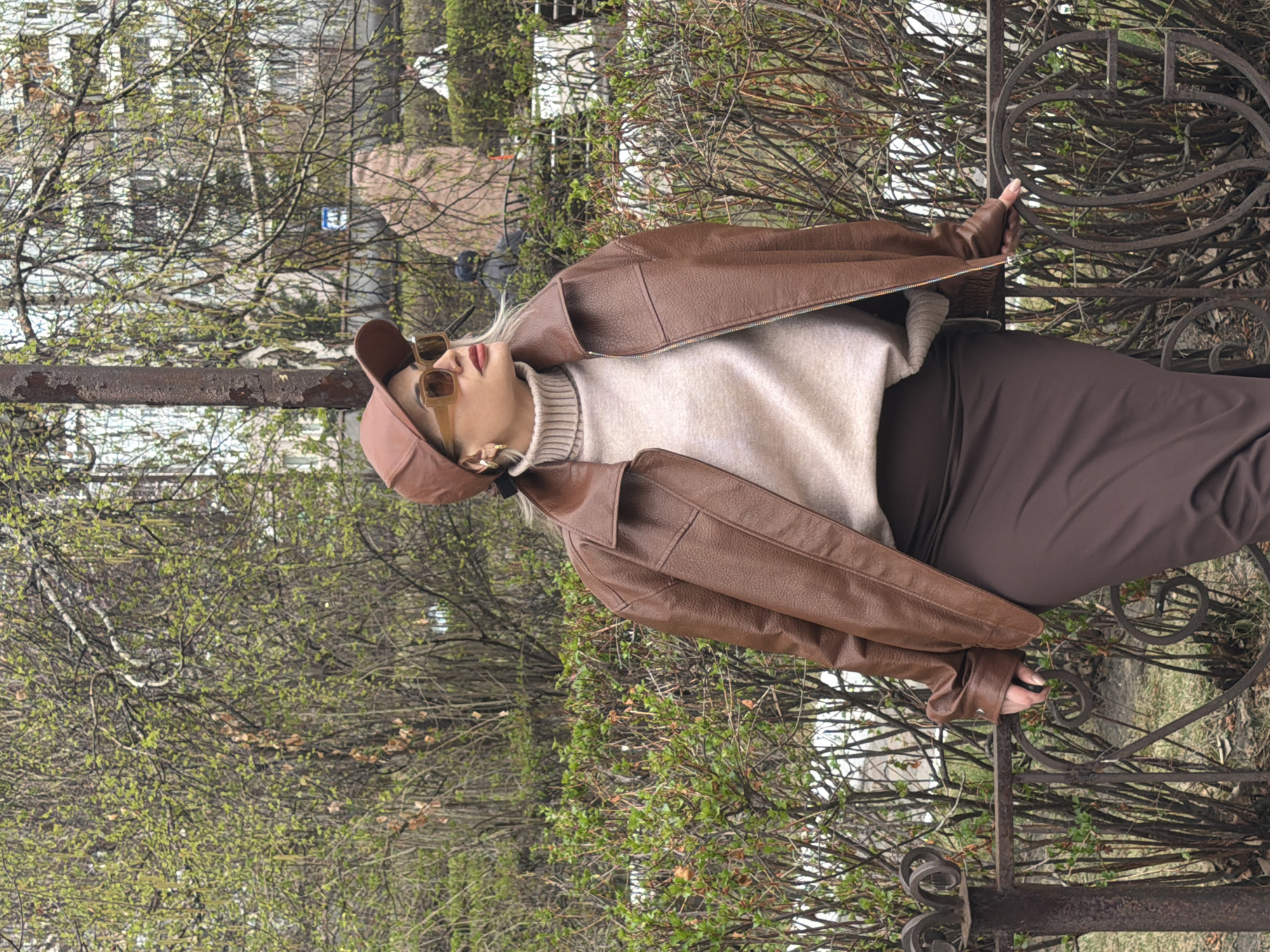 A fair-skinned person wearing a brown cap, sunglasses, brown leather jacket, a cream-colored turtleneck sweater, and a brown skirt stands outside in front of a wrought-iron fence. The person is positioned sideways. A tree and shrubbery are behind the fence. Buildings are visible in the background, along with street signs. The image was taken in Krasnoyarsk, Russia.
