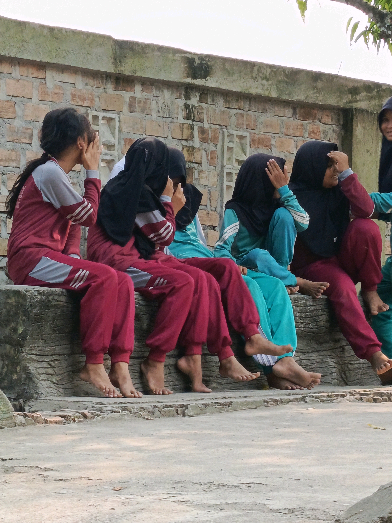 Five young individuals are seated on a rough stone ledge in front of a brick wall in Karya Dadi, Indonesia. They are wearing athletic-style clothing. The three individuals on the left are wearing maroon and grey tracksuits, while the two individuals to their right are wearing teal and maroon tracksuits, respectively. All five individuals have their hands covering their faces, obscuring their expressions. The background consists of a weathered brick wall and a blurry tree branch against a bright sky. The ground in front of the ledge is paved with concrete.