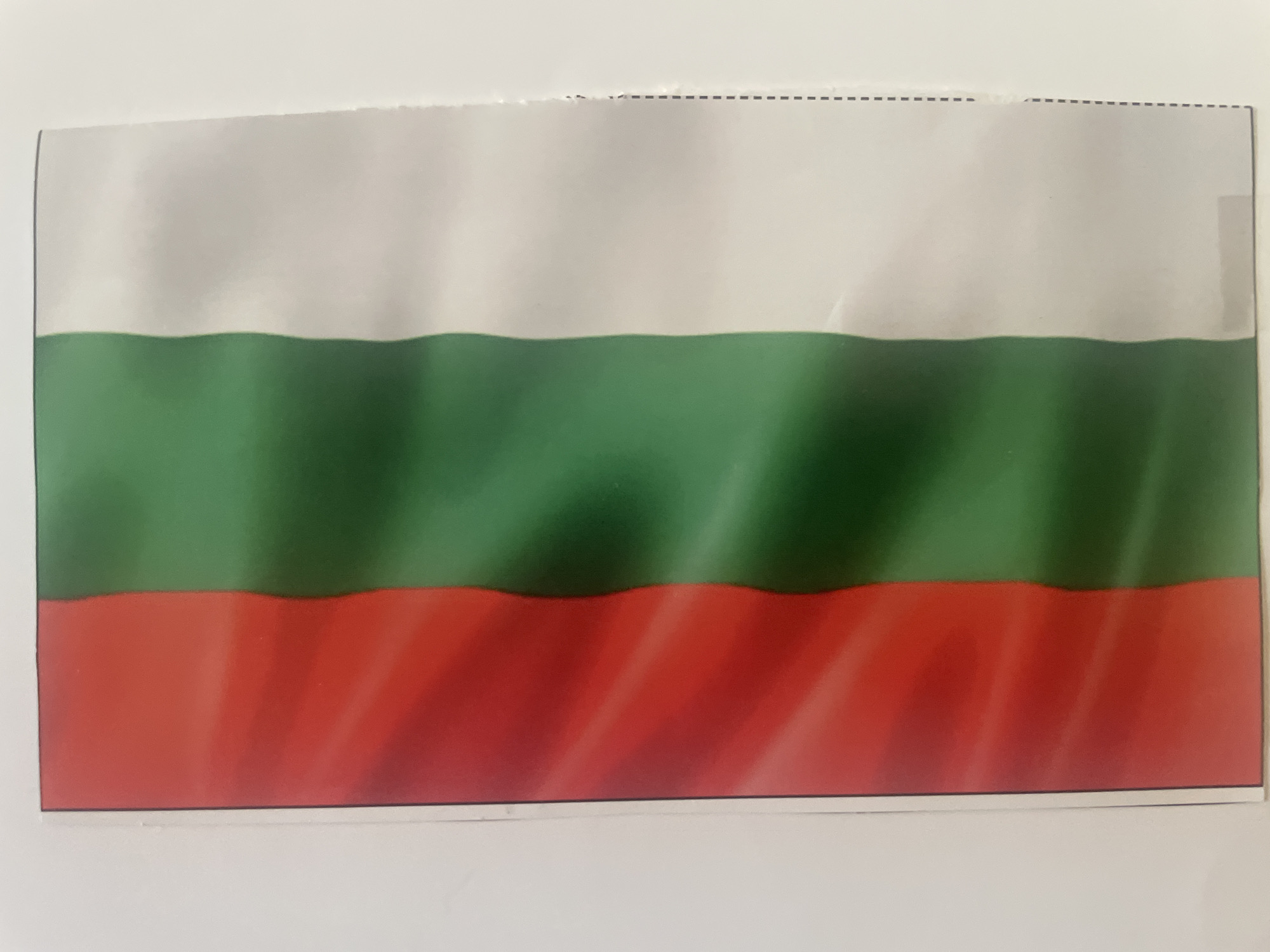 The image displays a printed representation of the national flag of Bulgaria, featuring three horizontal stripes. From top to bottom, the stripes are white, green, and red. The flag exhibits subtle visual textures, such as undulations and slight creases, which create the impression of a fabric flag, though it appears to be printed on paper.
The top edge of the flag shows an irregular, somewhat torn or jagged appearance, accompanied by a dashed line, suggesting it might have been separated from a larger document or sheet along a perforation. The flag is positioned against a clean, plain white background, which emphasizes its colors and design. There are no individuals or discernible activities taking place; the image is a static portrayal of the flag itself. The context of Yana, Bulgaria, aligns with the visual identification of the flag. No discernible text, beyond the aforementioned dashed line, is present.