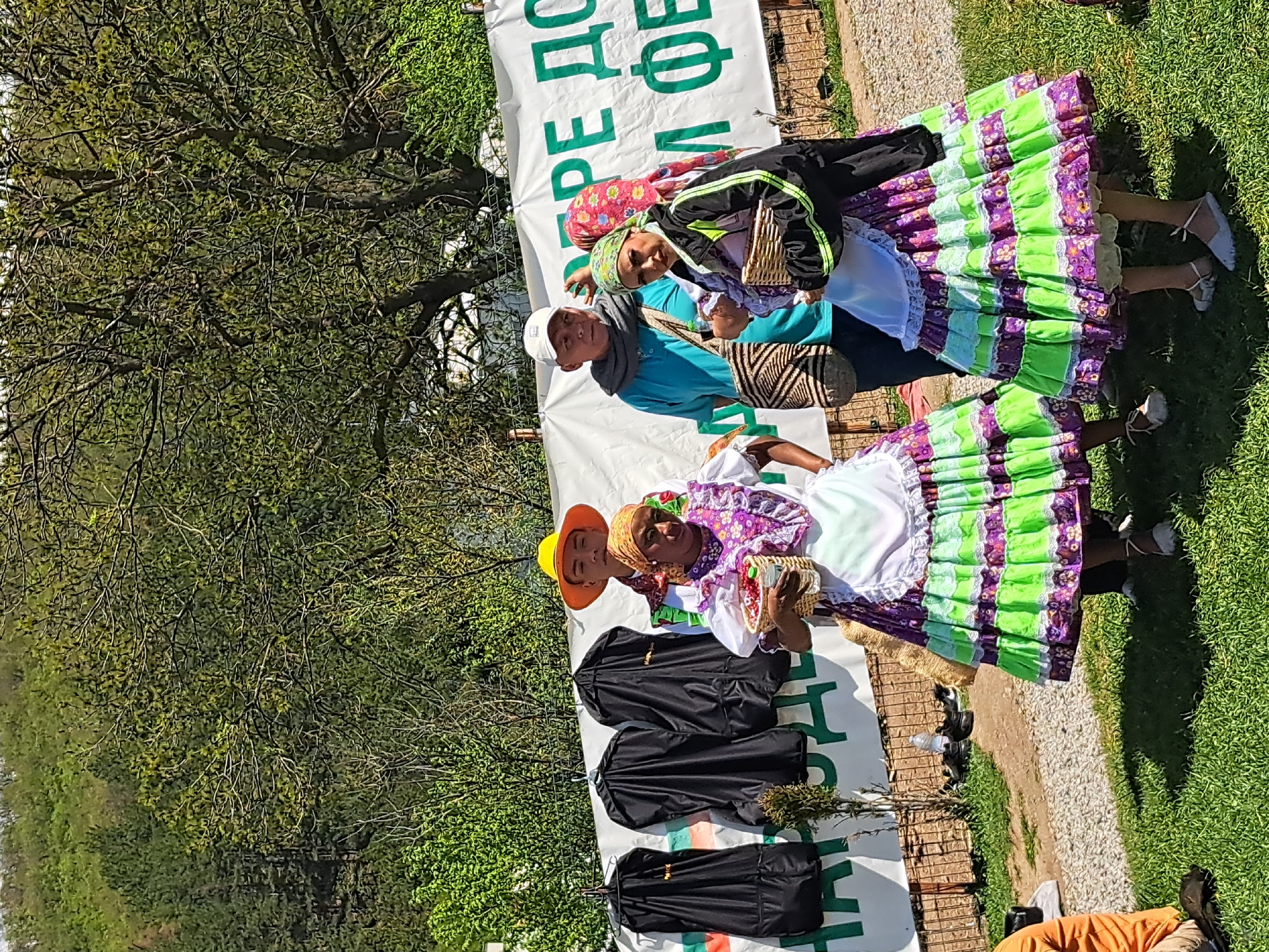 The image depicts three people in traditional clothing standing in front of a banner with text in what appears to be Bulgarian. The people are positioned on a grassy area next to a gravel path. A tree is visible in the background. In front of the people is a white banner. On the banner are black garment bags, with additional objects laying next to the bags. The location appears to be outdoors during the daytime. The city and specific location in Sofia, Bulgaria, cannot be determined with certainty from the visual information provided.