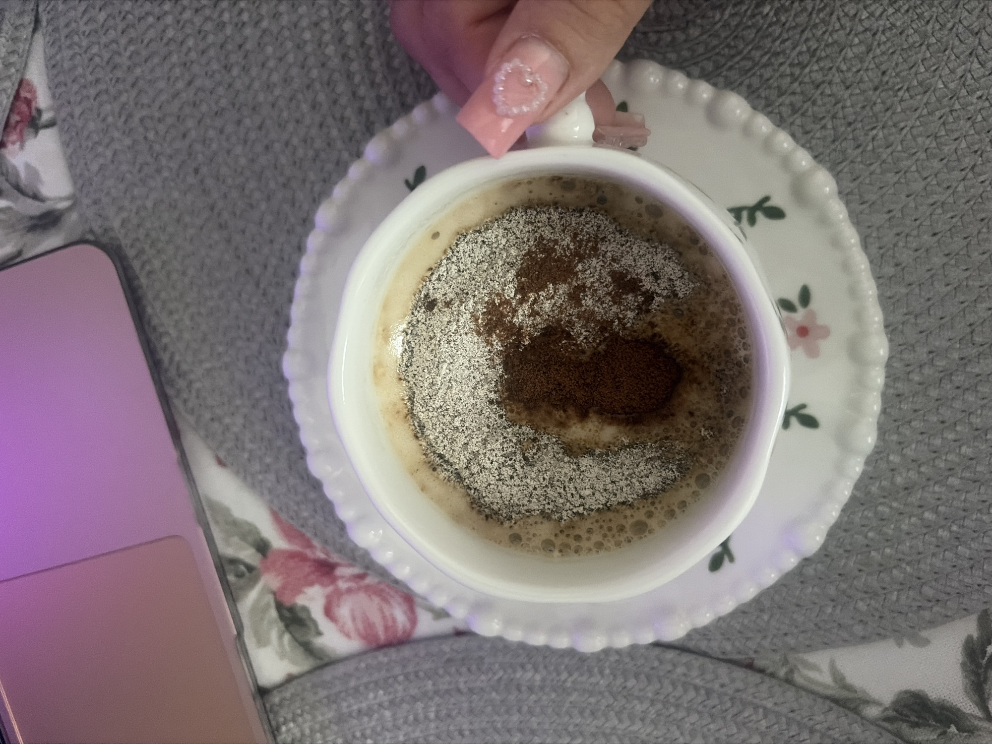 An overhead, close-up shot shows a woman's manicured hand, with light pink polish and a subtle white design on the ring finger, reaching towards a white teacup. The teacup is filled with a frothy beverage topped with a sprinkle of light brown powder and a darker brown powder in the center. The cup rests on a white saucer with a scalloped edge and a delicate floral pattern of pink flowers and green leaves. The background is a textured gray blanket layered over a floral-patterned fabric. A portion of a rose-gold laptop with a purple screen glow is visible in the upper left corner. The scene is set indoors, with no discernible urban or natural environment.