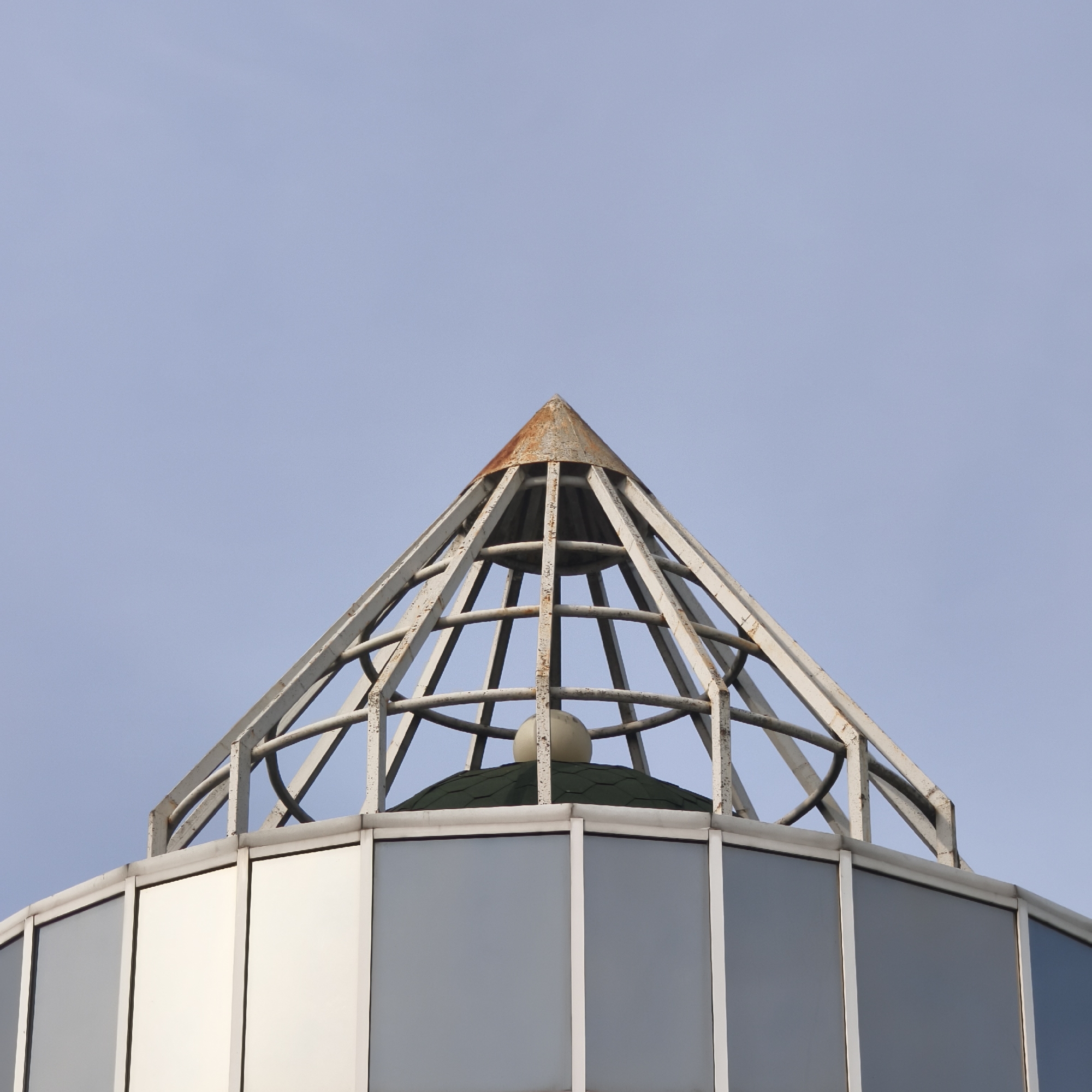 The image depicts the top of a building with a pyramidal metal framework structure. This structure surmounts a lower level with a glass-paneled exterior. At the apex of the pyramid is rusted metal. Inside the pyramid is a green-tiled dome topped with a light-colored sphere. The background is a pale blue sky. The location is in the city of Hrodna, Belarus.