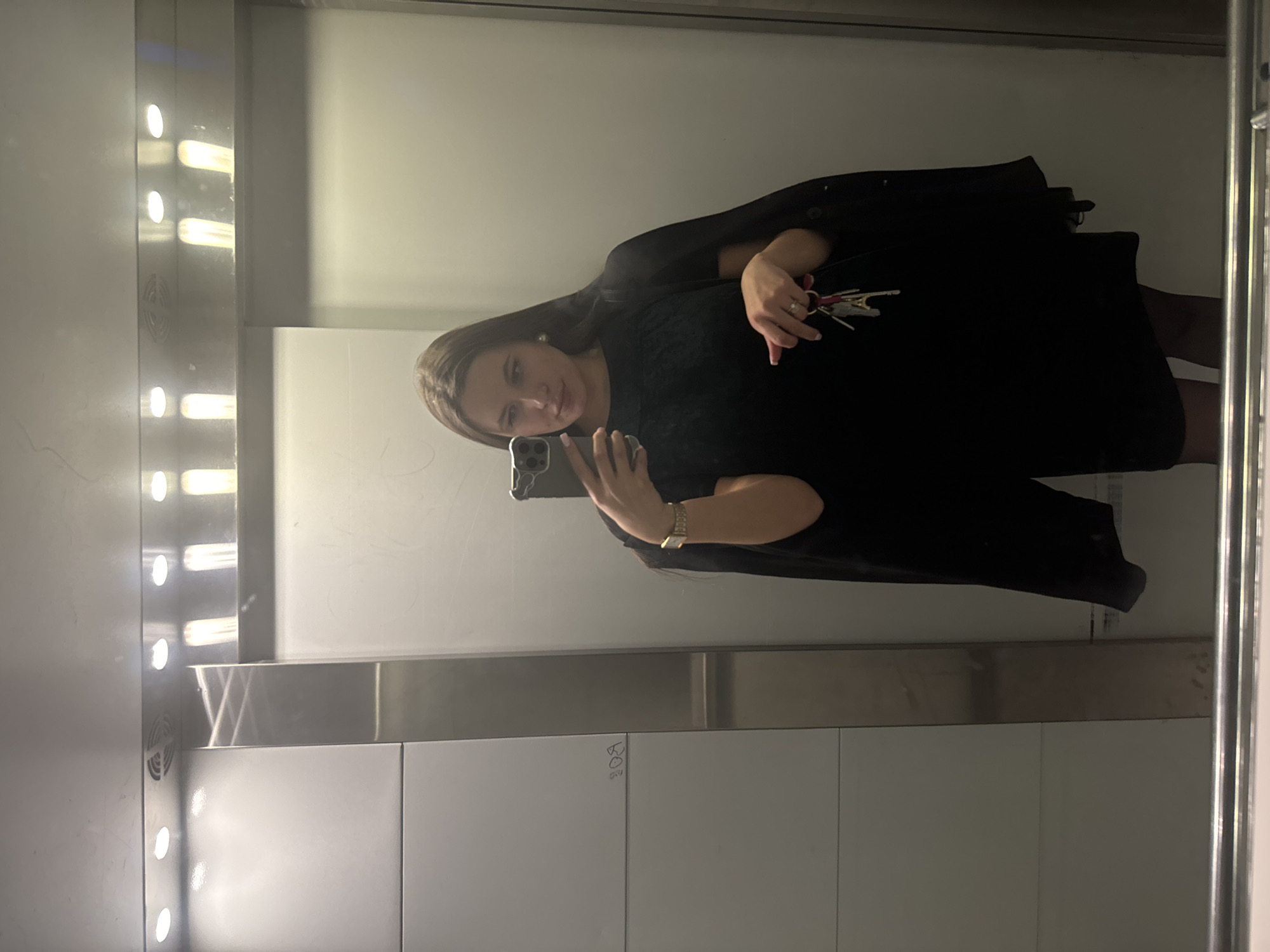 A female stands in an elevator, holding keys in her right hand and a smartphone in her left. The elevator is illuminated by a vertical row of lights. The walls are a combination of white and metallic panels. There is some graffiti on the wall. The location is in the city of Odintsovo, Russia.