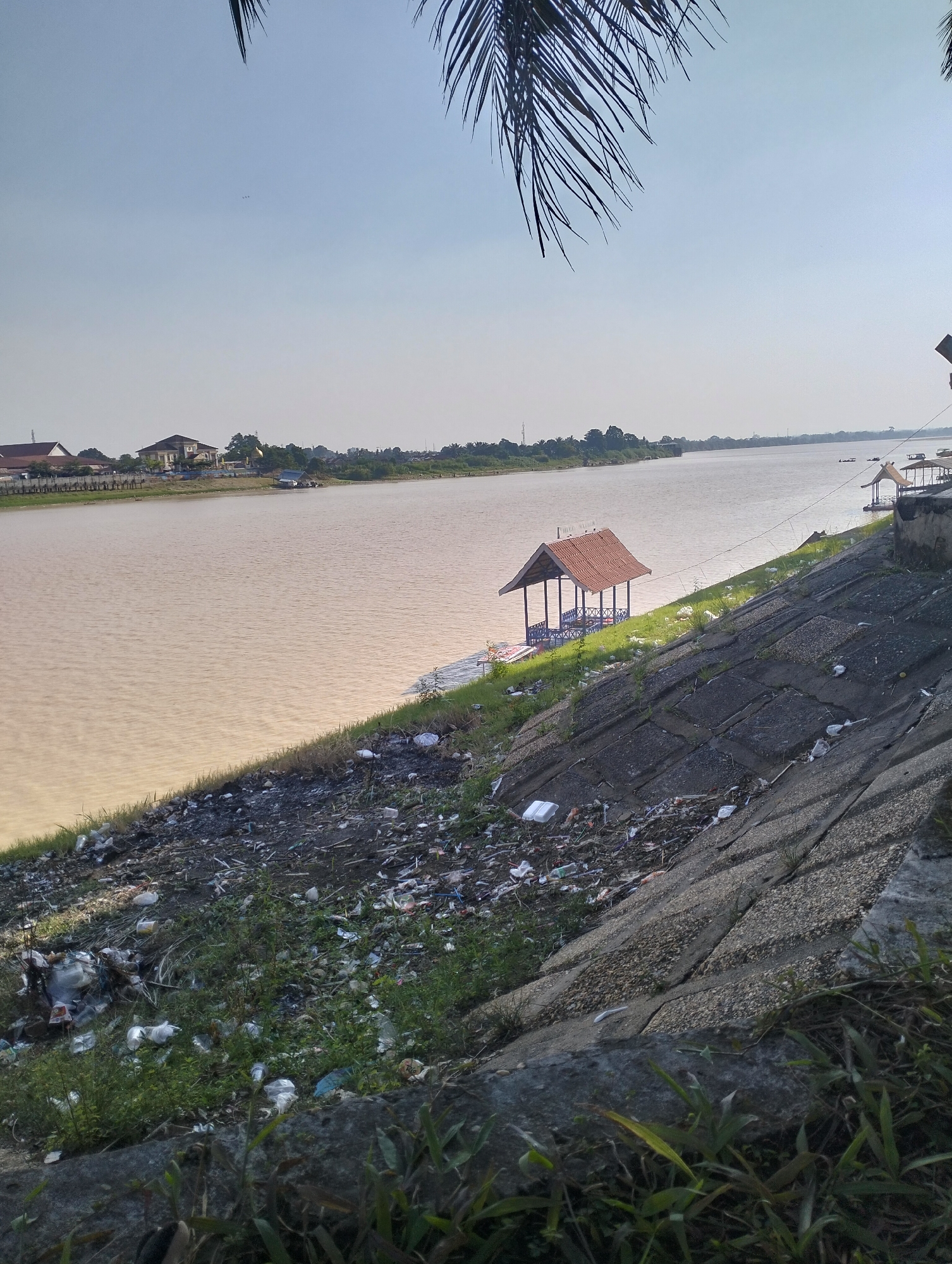 The image depicts a wide, muddy river flowing through a rural area, likely on the outskirts of Jambi City, Indonesia. The foreground is dominated by a steeply sloped embankment covered in debris and overgrown vegetation, with a concrete structure visible at the bottom. Scattered trash, including plastic bottles and bags, litters the ground.
On the riverbank, a small, open-sided pavilion with a red tiled roof stands on stilts, accessible by a wooden walkway. Several other structures, possibly floating platforms or small boats, are visible further down the river. In the distance, a cluster of buildings, including houses and what appears to be a mosque with a golden dome, line the opposite bank. The sky is clear and bright, suggesting a sunny day. The overall scene conveys a sense of neglect and environmental pollution along the riverfront.
