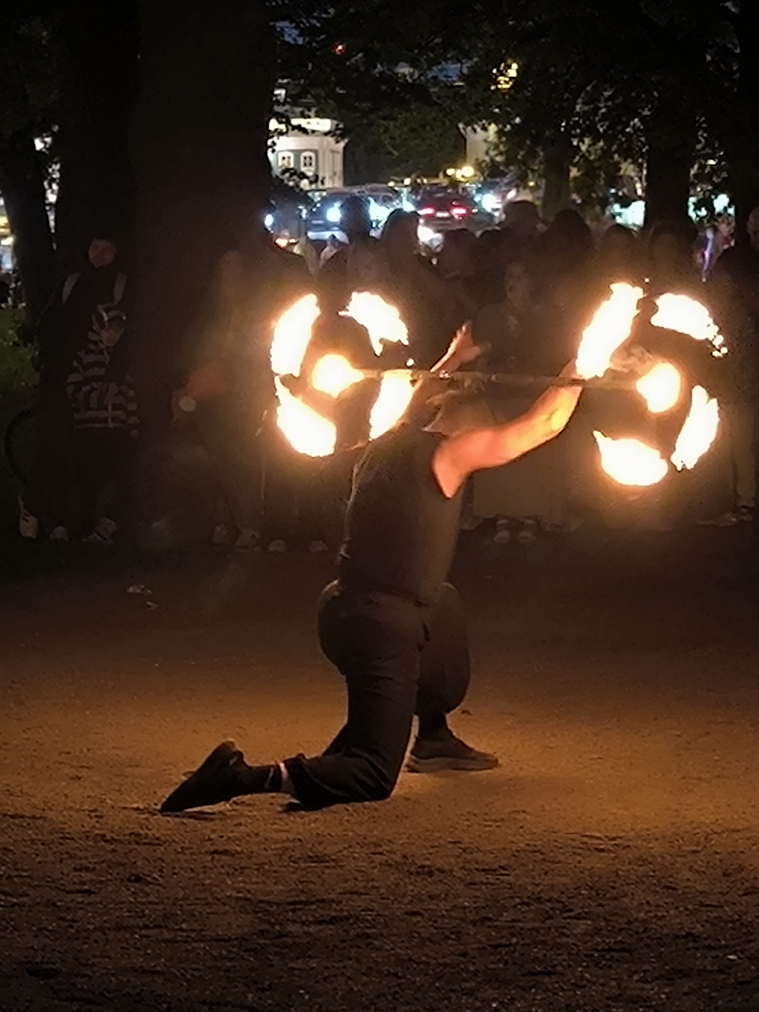 A full-length, eye-level, low-light photograph captures a nighttime fire performance in an outdoor urban or semi-urban environment in Unknown, Unknown. In the foreground, a single individual, wearing dark short-sleeved clothing, dark trousers, and dark athletic shoes, kneels on their right knee with their left foot flat on the ground. They are performing with a long, thin staff held horizontally across their upper back and shoulders, gripped by both hands. The staff's ends are ignited at multiple points, creating two distinct, bright orange-yellow fiery halos or spirals of light, indicating motion. The flames intensely illuminate the performer's arms and cast a warm glow on the immediate ground.
In the mid-ground, numerous indistinct, dark silhouettes of spectators are visible, positioned behind the performer. The background consists of dark tree silhouettes against a dimly lit cityscape. Distant artificial light sources include vehicle headlights and taillights on a visible street, and illuminated windows of various buildings, including a prominent white structure with dark rectangular openings to the left of center. The ground immediately surrounding the performer is a dark, granular surface, likely dirt or gravel, brightly lit by the fire. The scene is predominantly dark, with the fire acting as the primary light source and focal point.