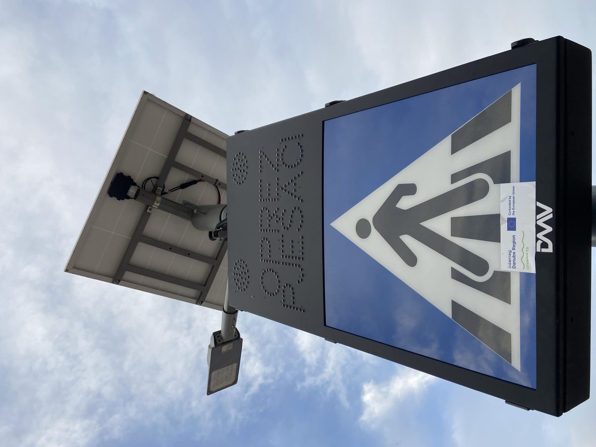 The image depicts a solar-powered pedestrian crossing sign in Budva, Montenegro. The sign is mounted on a gray pole. A solar panel is attached to the top of the pole. The sign itself is rectangular with a blue background and a white triangle containing a black pedestrian silhouette. To the side of the sign, holes in the pole spell the word "PEŠAČKI." A sticker is attached to the pedestrian crossing sign that contains the EU flag and the text "Interreg Danube Region CITYWALK 2.0." A streetlight is attached to the pole below the solar panel. The background is a sky with clouds.