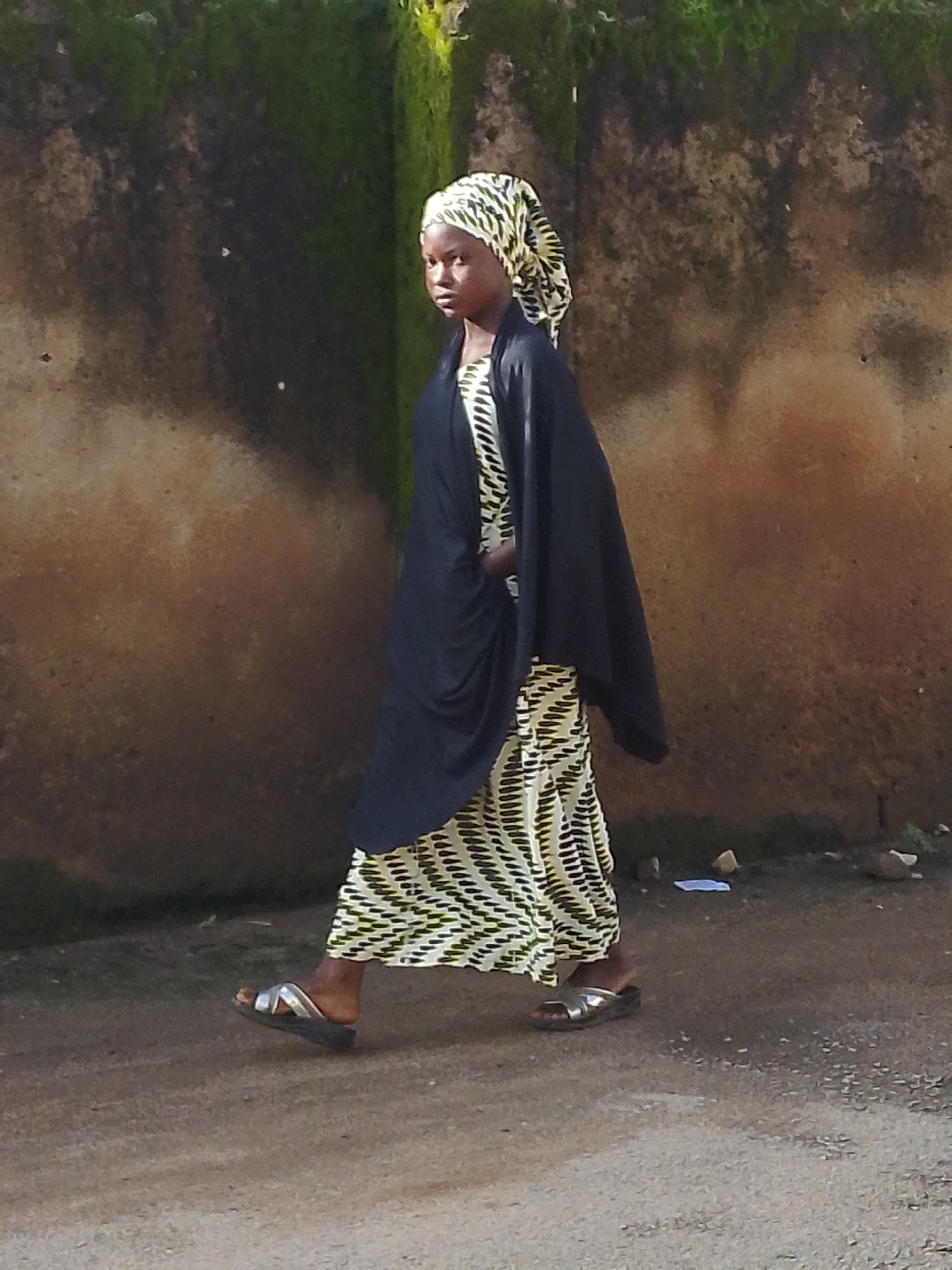A young person wearing a black shawl over a patterned dress and a headscarf walks along a dirt path next to a textured wall. The wall, covered in patches of green moss, appears weathered and shows a gradient of brown tones. The ground is a dusty, light brown with small pebbles and debris scattered on its surface. The person is wearing silver sandals. The scene suggests an outdoor environment in a city like Jos, Nigeria.