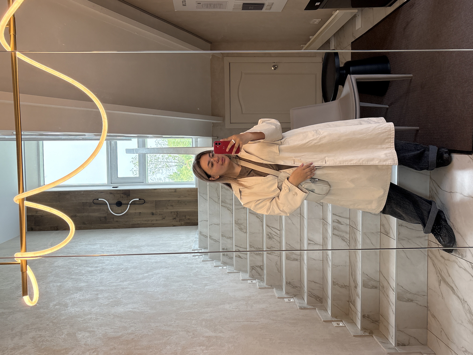 A young woman is taking a selfie in a mirror on a staircase. The staircase is made of white marble with grey veining. The background features a window, a light fixture, and elements of a modern interior design, possibly in a building located in Khabarovsk, Russia. The woman is wearing a light-colored jacket, dark pants, and glasses, and is holding a red smartphone. There is a light-colored purse hanging at her side. The mirror reflects the staircase, the woman, and elements of the room behind her, including a curved modern light fixture.
