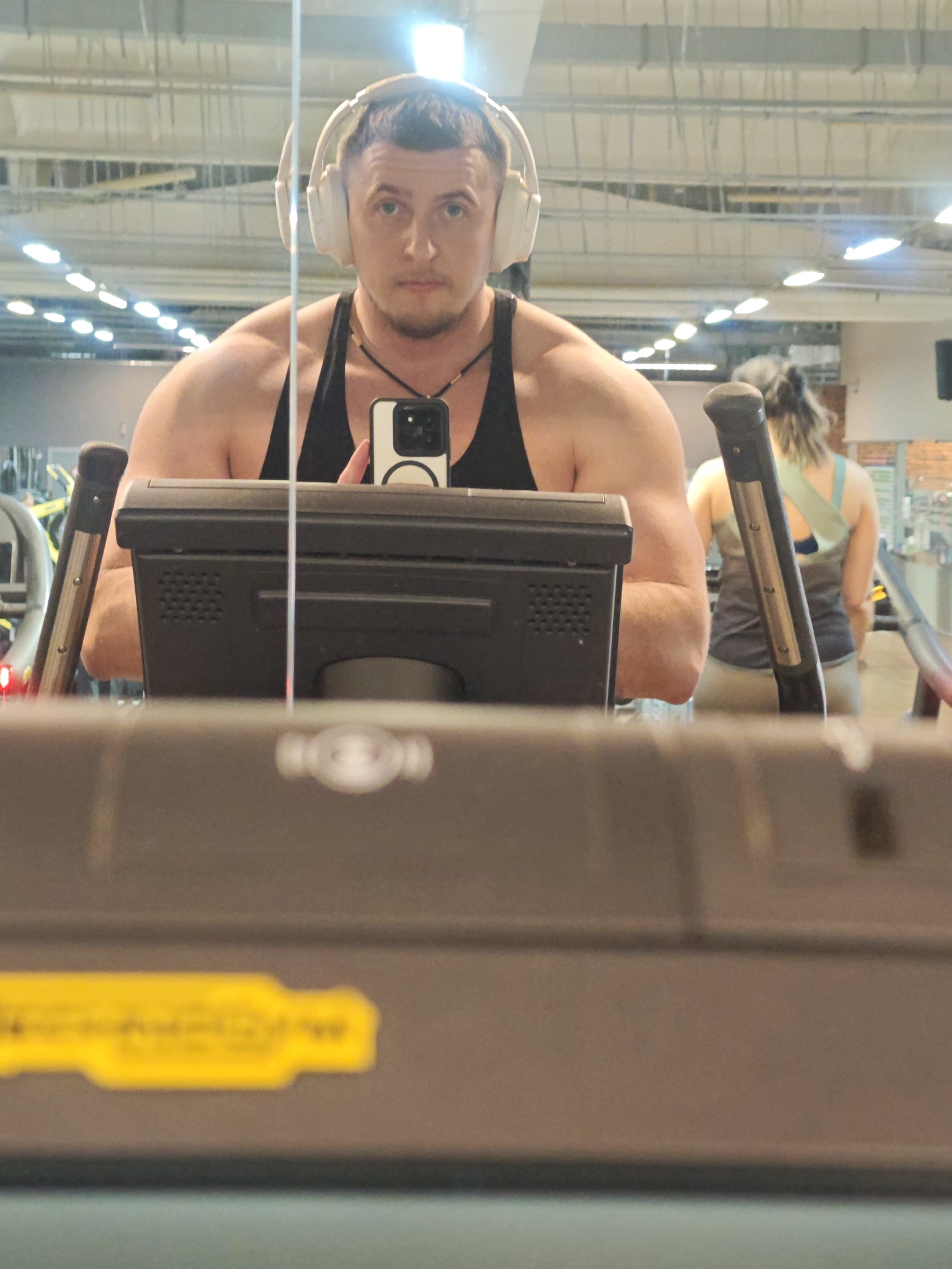 A man is taking a selfie in a mirror at a gym. He is wearing a black tank top and white headphones. He's holding a phone in front of his face. In the background is another person on a treadmill. The gym appears to be indoors, with fluorescent lighting and exposed ductwork. The image does not provide sufficient information to confirm it was taken in the city of Chaikovsky, Russia.