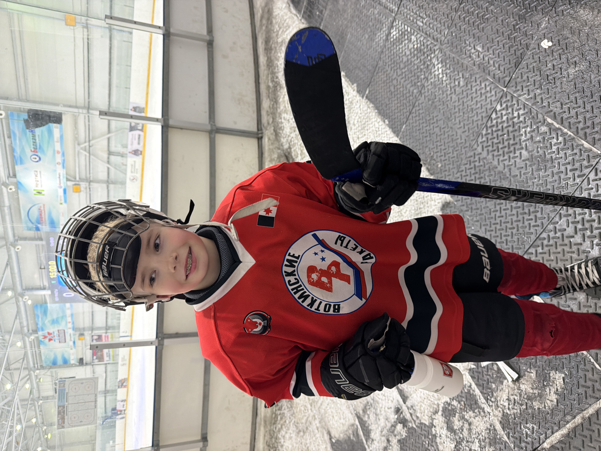 A young boy stands on a textured, gray metal floor. He's wearing a hockey helmet, a red hockey jersey with black and white accents, black gloves, and red pants. He is holding a hockey stick in his right hand and a white water bottle in his left. The jersey has a circular patch on the front with the words "ВОТКИНСКИЕ" (Votkinskiye) and "РАКЕТЫ" (Rakety) in Russian, and features an emblem with the Cyrillic letters "ВР". The jersey also has a small Canadian flag patch on the shoulder. The background shows an ice rink with advertising banners and a scoreboard. Considering the jersey inscription "Votkinskiye Rakety" (Votkinsk Rockets) and the Cyrillic text, the location is likely an ice rink in Votkinsk, Russia. The boy is smiling, revealing braces on his teeth.