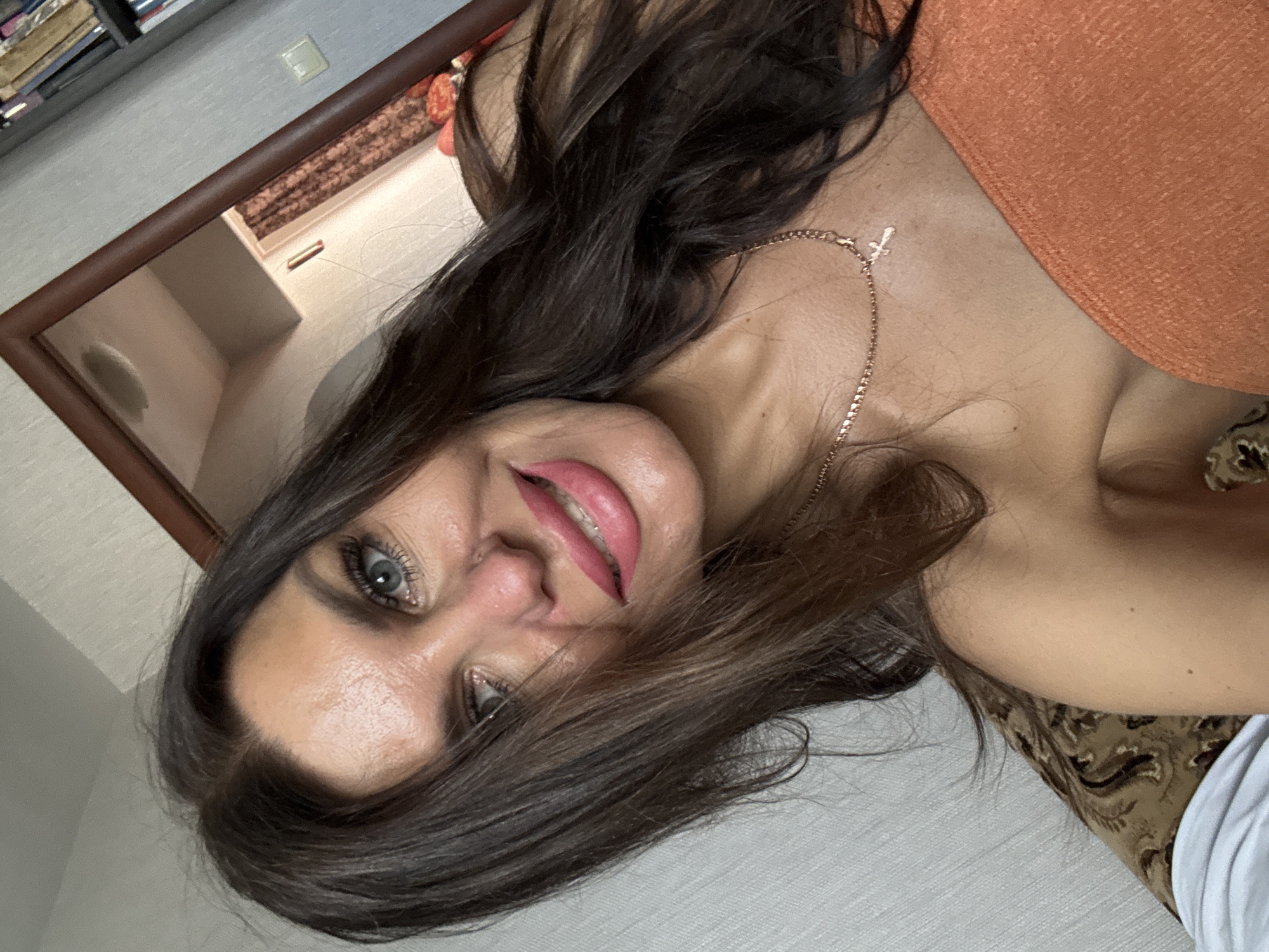 The image shows a selfie of a woman with long, dark brown hair, green eyes, and pink lipstick. She is wearing an orange strapless top and a rose gold necklace with a cross pendant. She is indoors, possibly in a bedroom, with a mirror in the background reflecting a room with curtains. There is a bookshelf visible in the upper right corner of the image. The environment appears to be domestic. There is no indication of the specific city or location within Dubovaya, Russia.