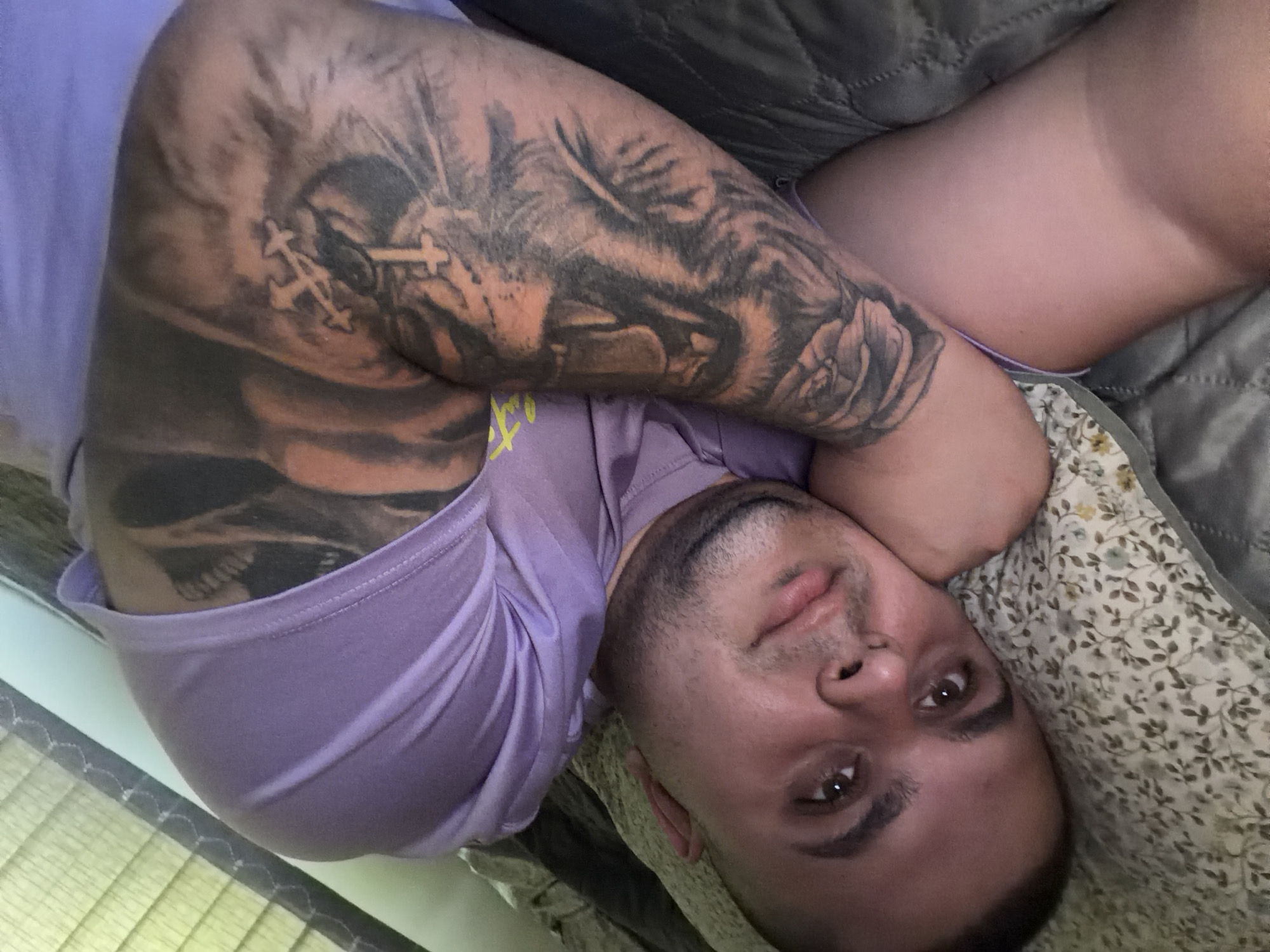 A man is captured in a close-up shot, lying down indoors, likely in a bedroom setting. He has short, dark hair, light brown eyes, and stubble on his face, looking directly at the camera with a neutral expression. He is wearing a light purple short-sleeved t-shirt.
His right arm, which is bent and positioned beneath his head, is adorned with a detailed sleeve tattoo. Key elements visible in the tattoo include multiple crosses, a prominent depiction of a large animal (possibly a lion or wolf) on the upper arm, a detailed rose near the elbow, and what appears to be a skull or skeletal imagery further down the arm.
He is resting on a floral-patterned pillow. A grey quilted blanket or bedspread is visible behind him and partially covering his lower body. To the left, a window with horizontal blinds can be seen in the background. The lighting is soft and even, consistent with an indoor environment. There is no visible text in the image. The location context provided is Shumen, Bulgaria, though no visual cues within the image confirm this.