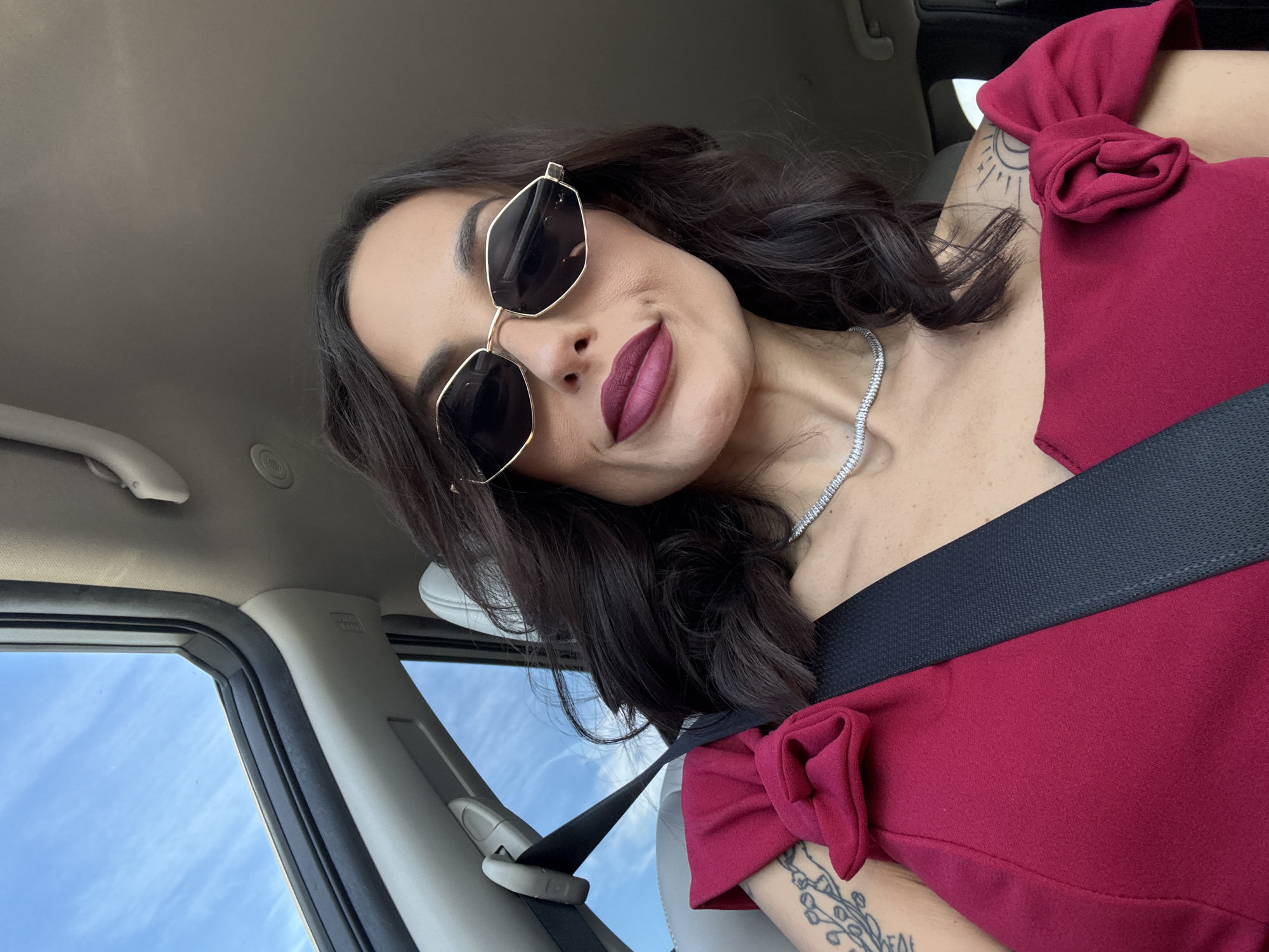 A woman with dark hair and sunglasses is inside a car. She is wearing a red dress with bow-like details on the shoulders and a diamond necklace. A seatbelt is fastened across her body. Tattoos are visible on her arms. The interior of the car is visible with parts of the door and window frame shown. The background visible through the car window indicates a bright, sunny day with a blue sky. There is no indication of Yasen, Bulgaria.