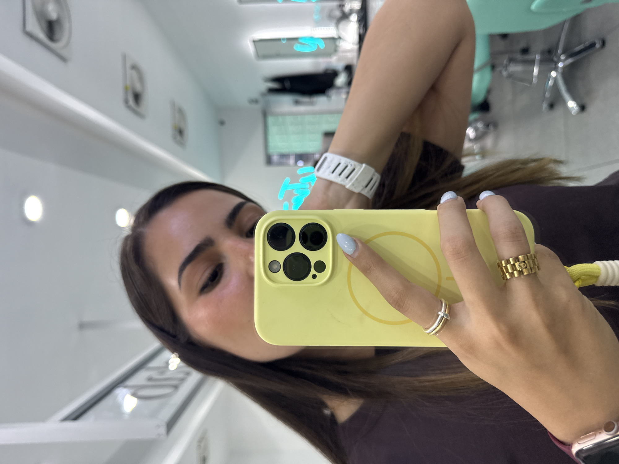 A person is holding a yellow-cased iPhone up in front of their face in a mirror selfie. The person has brown hair, brown eyes and is wearing two rings on the hand holding the phone. The person is also wearing a white smartwatch on their left wrist. The location seems to be a commercial space, based on the interior decor. Without additional evidence, it is not possible to confirm the location as San Bernardino Tlaxcalancingo, Mexico.