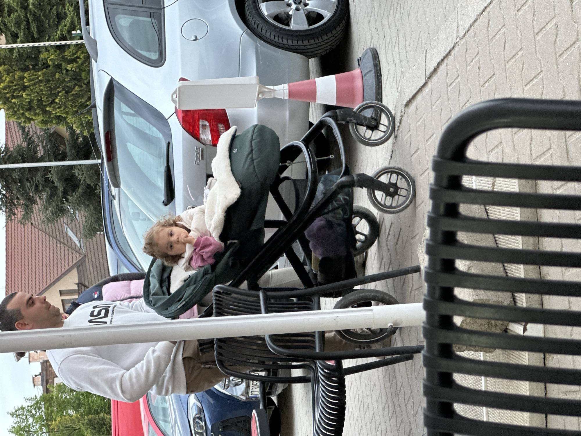 The image captures an outdoor urban scene, likely in Plovdiv, Bulgaria, featuring a man and a young child next to parked cars. The scene is oriented vertically, implying the original photo was taken in portrait mode or rotated.
In the foreground, a man with short dark hair is seated, facing left, looking down towards a child. He wears a white sweatshirt with "NASA" clearly printed on the front, along with smaller text underneath. He is wearing brown pants. Partially visible in the lower right foreground are black metal slatted chairs or benches. A white horizontal bar or pole runs across the mid-ground.
To the man's right and slightly in front of him, a young child with curly light brown hair is seated in a dark green baby stroller with a white fleece lining. The child is wearing a light-colored jacket and holds a pink object near her mouth, looking towards the camera with a neutral expression.
Behind the man and child, several cars are parked. A prominent silver hatchback, identifiable as a "Fabia" (Skoda Fabia) from text on its rear, is parked with its rear towards the viewer. Next to it, a red and white striped traffic cone with a white rectangular top stands on the pavement. Further back, parts of a dark blue car and a red car are visible. The blue car has a license plate partially visible as "CT 3053", which corresponds to the Plovdiv region in Bulgaria.
The ground is paved with patterned concrete tiles. In the background, green trees and parts of buildings with red-tiled roofs suggest a residential or mixed-use area. The lighting appears soft, indicating an overcast day or perhaps late afternoon. No specific activity beyond casual waiting or observation seems to be taking place.
