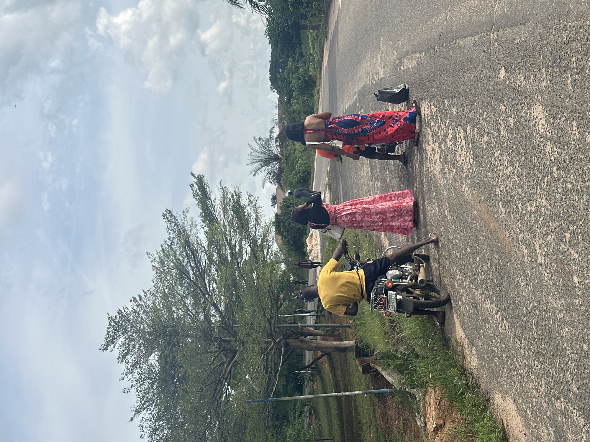 The image shows a man on a motorcycle stopped on a paved road in Ihiagwa, Nigeria. Two women in long dresses stand next to the motorcycle. The background includes trees and sky. The motorcycle's license plate reads "EHH-116QT". A black plastic bag sits on the road near the women.