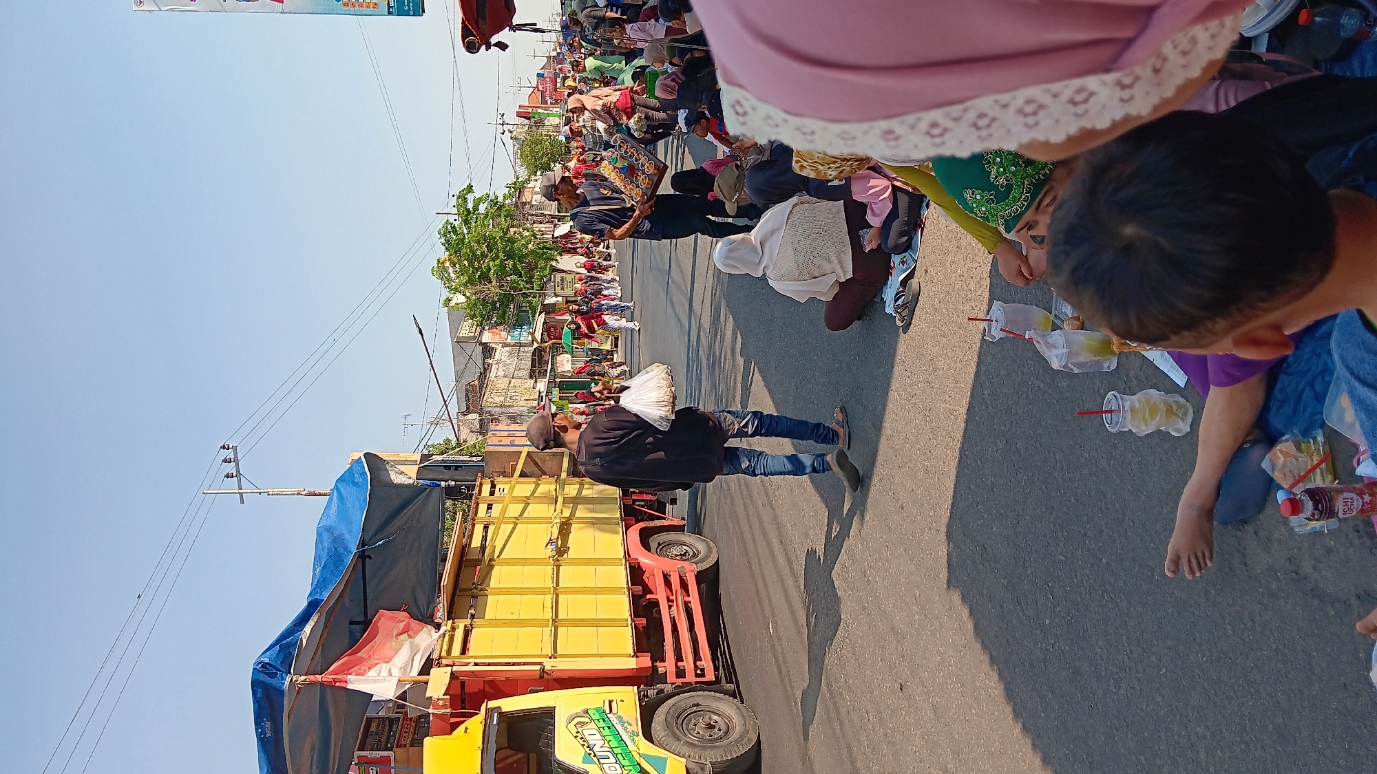 In Tanjung Anom, Indonesia, a street scene unfolds under a clear blue sky with a bright sun casting long shadows. The foreground is occupied by a group of people, some seated on blue mats, others partially visible from behind. A child's head and shoulder are prominent in the lower right corner. Several clear plastic cups with red straws, some containing a yellow-green liquid and ice, are scattered among them. A bottle of "ICHIACHA" is also visible.
The middle ground features a yellow truck with a blue tarp covering its load, adorned with Indonesian flags. A man in a dark shirt and jeans walks away from the truck, carrying a white plastic bag filled with what appear to be food items. Another man stands in the street holding a tray of small, colorful cup cakes.
Beyond them, the street is lined with buildings and trees, suggesting a populated urban area. More people are scattered throughout the street, some walking, some gathered. The overall impression is a lively outdoor gathering or event taking place on a sunny day.