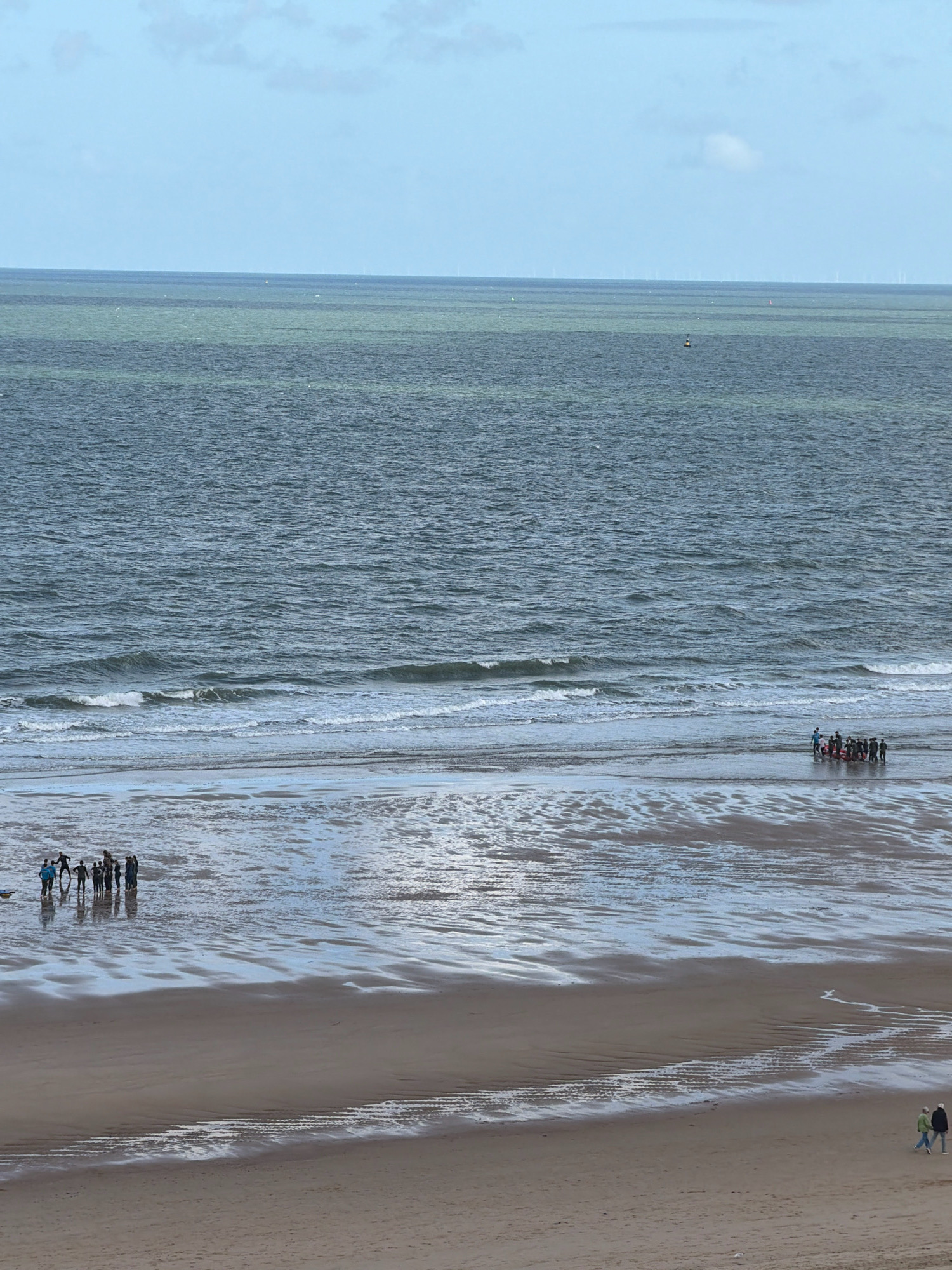 The image depicts a wide shot of the beach and ocean at Ostend, Belgium. The sky is a pale blue with scattered white clouds. The ocean is a deep blue-green, with gentle waves lapping at the shore. The beach is a mixture of sand and shallow water, reflecting the sky. In the foreground, two people are walking on the sand. Further down the beach, a group of approximately ten people are standing in the shallow water, some appearing to be engaged in a group activity. In the distance, on the horizon, several offshore wind turbines are visible. The overall scene suggests a calm, overcast day at the Belgian coast.