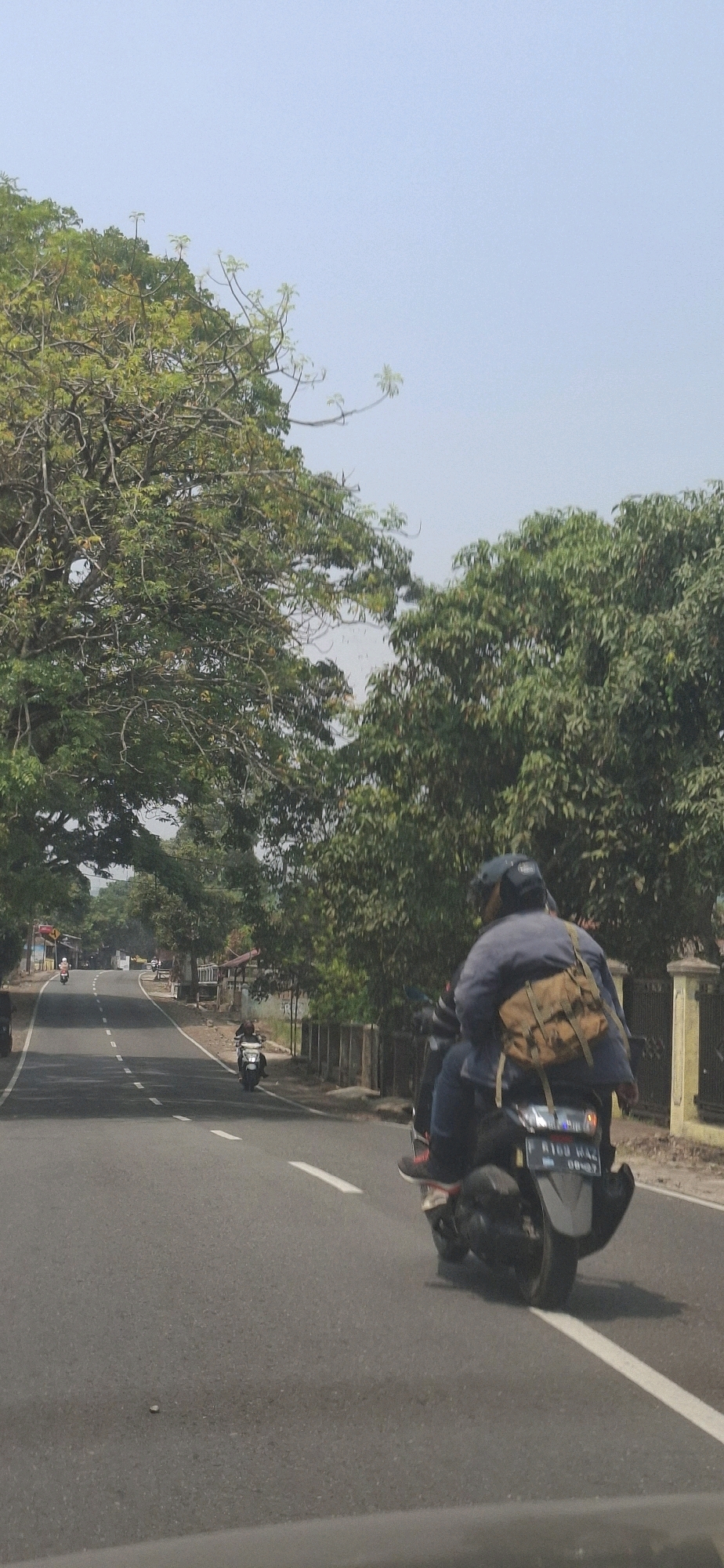 The image shows a street scene in Susukan, Indonesia, on a clear, sunny day. In the foreground, two people are riding on a scooter. The person in the back is wearing a blue jacket and a brown bag is slung over their shoulder. The scooter's license plate is partially visible. The road is paved with white lines marking the lanes. On either side of the road are lush green trees, with one large tree arching over the road in the left background. In the distance, other scooters and some buildings can be seen. The overall impression is of a typical day in a semi-urban or rural area.