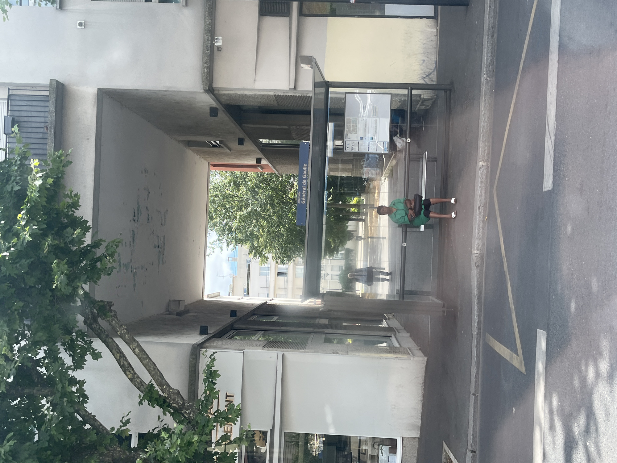 The image shows a street scene in Alfortville, France, specifically at a bus stop labeled "Général de Gaulle." In the foreground, there is a paved road with yellow markings. Behind the road is a sidewalk and a bus stop shelter. A person, identified as an adult, is seated on the bench inside the bus stop shelter, wearing a green shirt and dark pants, and white sandals. They are holding a dark bag on their lap. Reflected in the glass of the bus stop shelter, another person is visible walking away. The bus stop is situated in front of a multi-story building with balconies and windows. Overhanging the scene from the left is a large tree with green foliage. The background shows more buildings and trees under a bright sky.