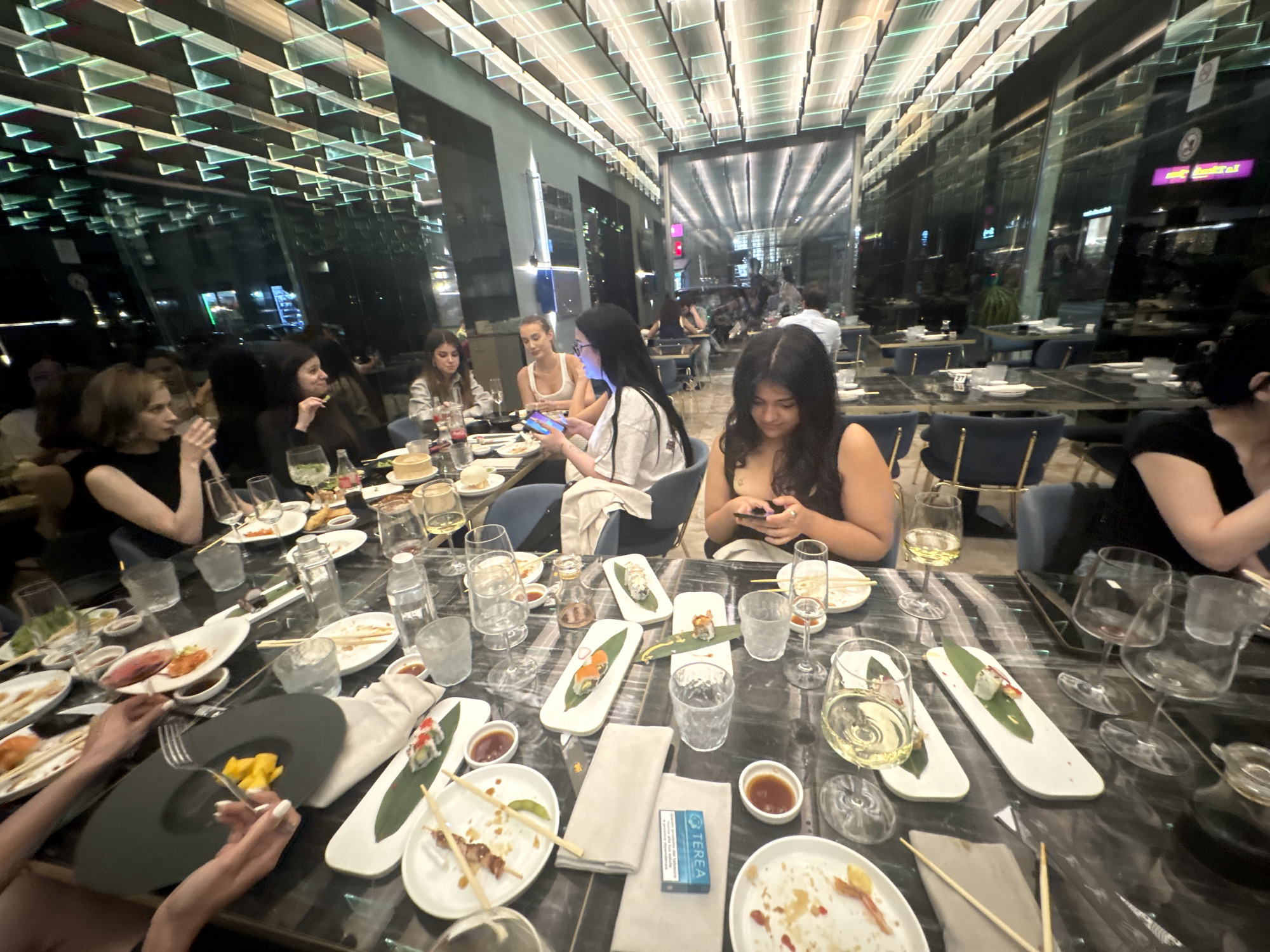 A group of individuals, appearing to be young adults, are seated around a large table in a restaurant. The table is laden with plates of sushi, chopsticks, various dishes, and drinks, suggesting a meal in progress. The individuals are engaged in eating, conversing, and one person is looking at their phone. The restaurant has a modern and upscale ambiance, characterized by a distinctive ceiling design with illuminated panels and reflective surfaces. The overall impression is that of a social gathering at a contemporary dining establishment, likely in the city of Milan, Italy, given the context provided.