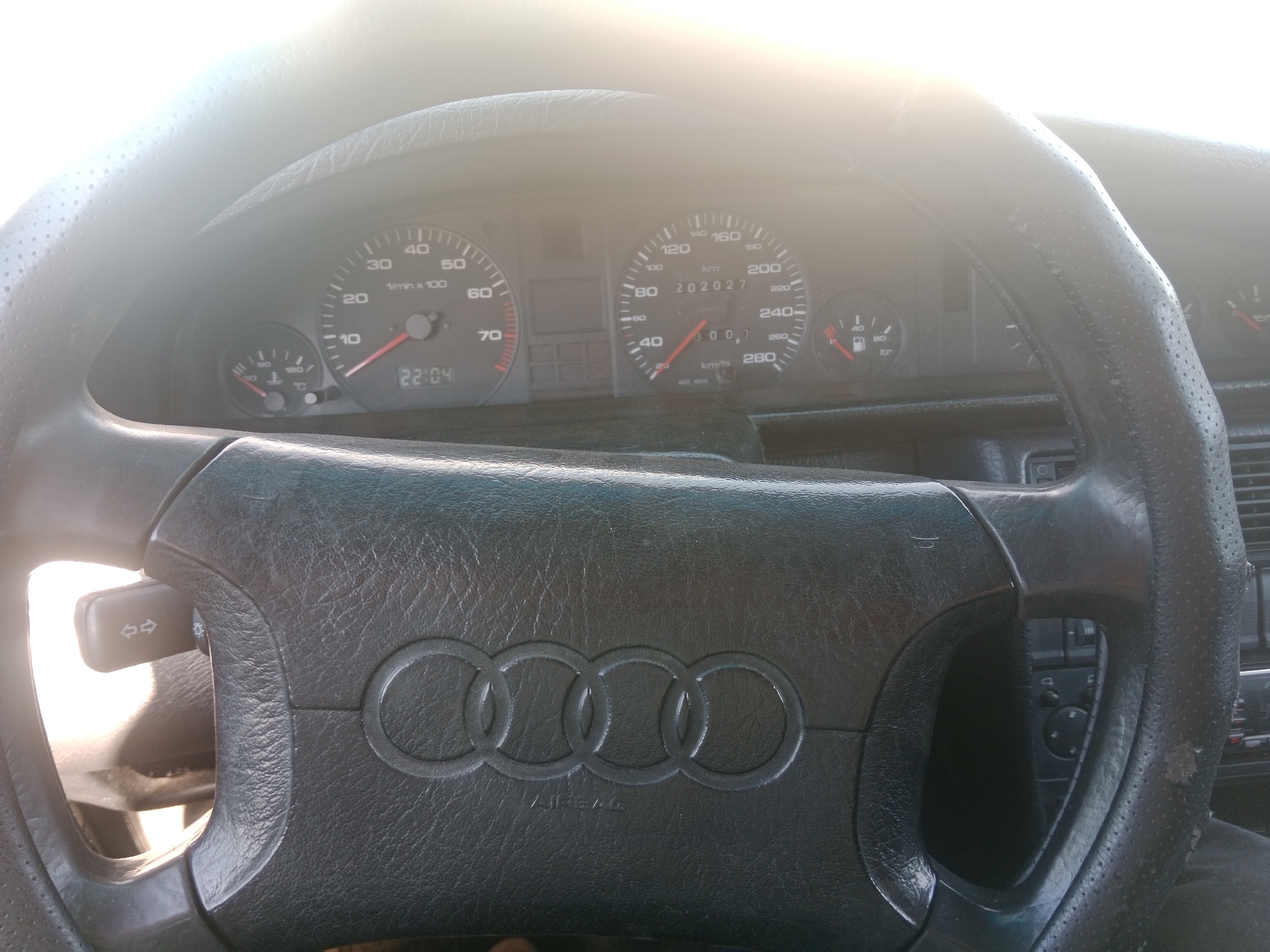 The image shows the interior of a car, focusing on the steering wheel and dashboard. The dashboard features various gauges, including a speedometer, tachometer, and fuel gauge. The speedometer shows a reading of 202,027 kilometers. The time displayed on the dashboard is 22:04. The steering wheel is black leather with the Audi logo prominently displayed in the center, along with the word "AIRBAG". A turn signal lever is visible on the left side of the steering column. The overall lighting suggests it is either early morning or late evening, with strong sunlight coming from the left side of the frame, causing some glare on the dashboard. The car is stationary, as indicated by the parked position of the needles on the gauges. The specific location in the city of Qobda, Kazakhstan is not identifiable from the visual information presented in the image.
