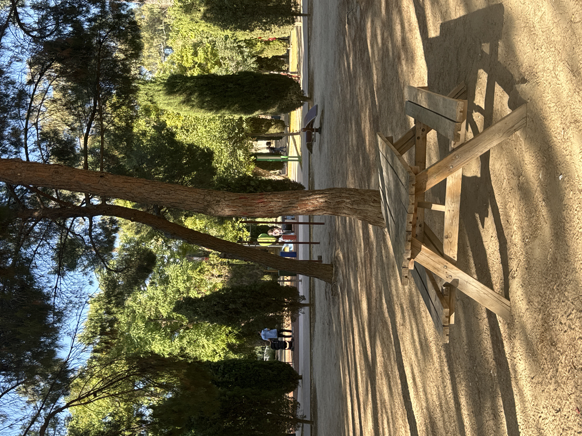 The image depicts a public park setting with abundant trees, a sandy ground surface, and dappled sunlight casting long shadows. In the foreground, a weathered wooden picnic table with attached benches is prominently featured. In the mid-ground, a person is visible walking a baby stroller, and further in the background, other individuals are present, along with playground equipment. The overall ambiance suggests a serene, outdoor recreational area. While the specific location within Murcia, Spain, is not explicitly identifiable from the image alone, the presence of such amenities is typical of urban parks found in cities like Murcia.