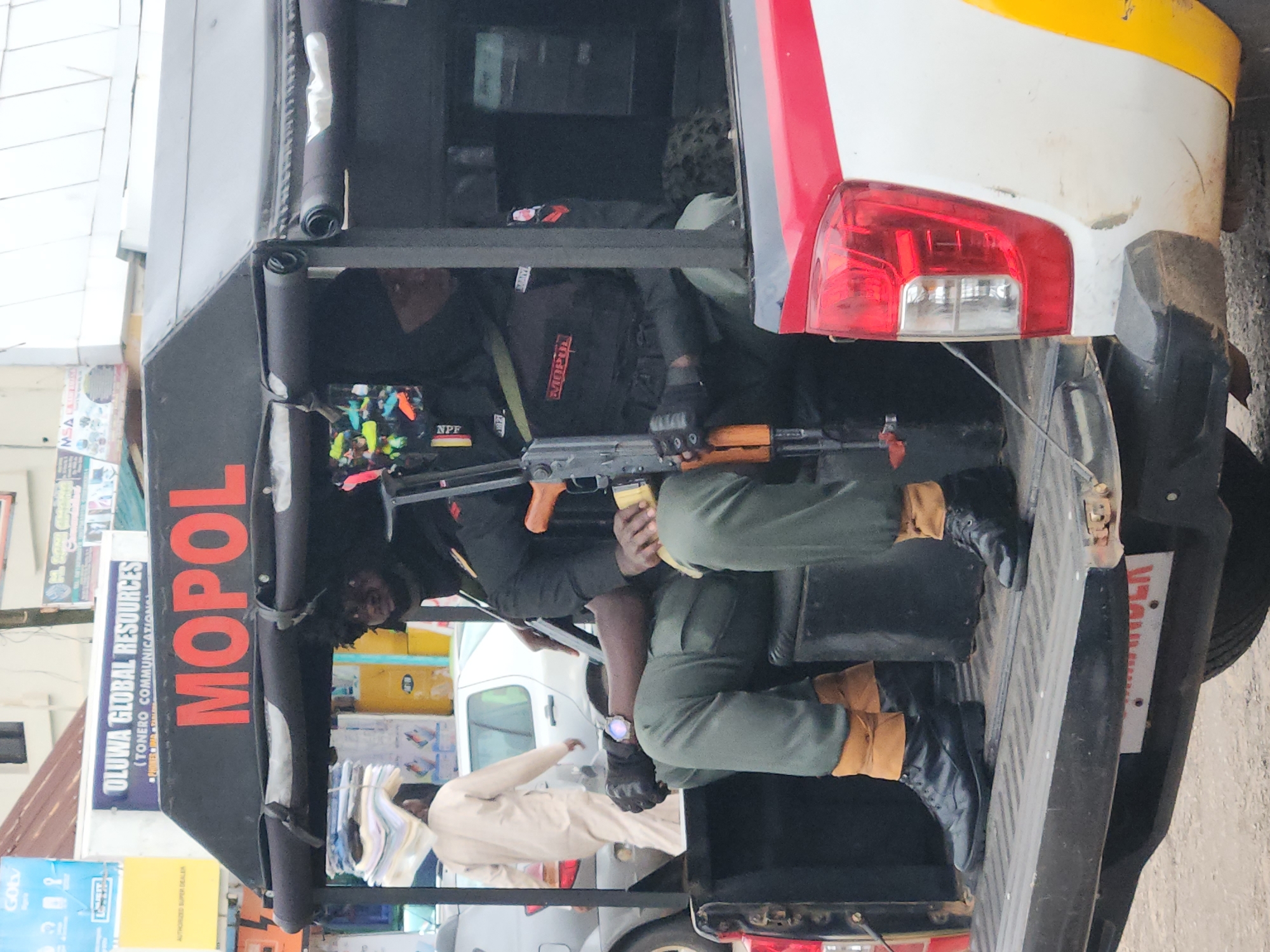 The image shows the rear of a pickup truck in a city, likely Jos, Nigeria, with two men in police uniforms sitting in the truck bed. The truck has a black canopy with "MOPOL" written in red lettering on the side. The men are wearing dark uniforms with patches on their sleeves, olive green pants, and black boots. One man is holding a rifle. In the background, there are buildings and other vehicles. One building has a sign that reads "OLUWA GLOBAL RESOURCES". A person in a light-colored robe is walking in the background carrying a stack of papers on their head.