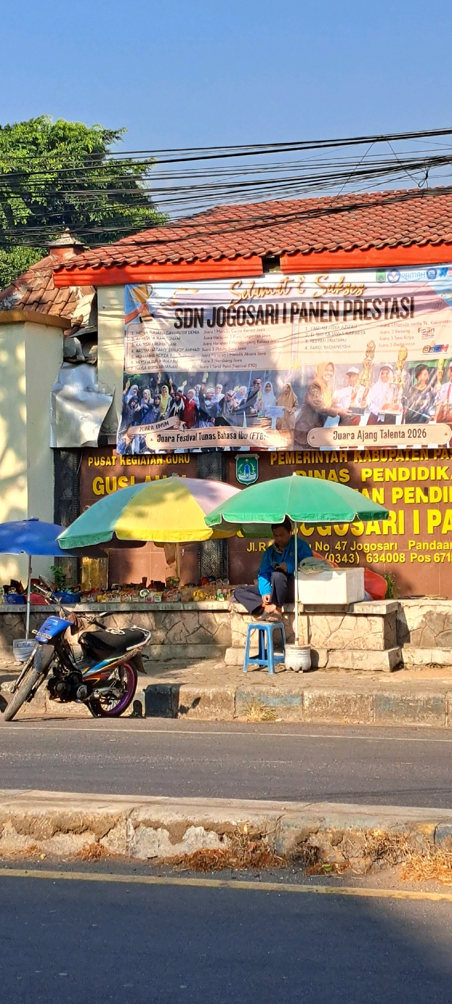 The image shows a street scene in Petungasri, Indonesia, under a clear blue sky. A banner is displayed on a building that reads "SDN JOGOSARI I PANEN PRESTASI" and lists various awards. Below the banner, a man is sitting on a blue stool under a green umbrella, looking at his phone. In front of him are some food items and a white cooler. To the left, a motorcycle is parked. Several umbrellas, a blue and a yellow one, are also visible. The building appears to be a school or community center, with a stone wall and a tiled roof. The area looks like a marketplace or roadside stall.
