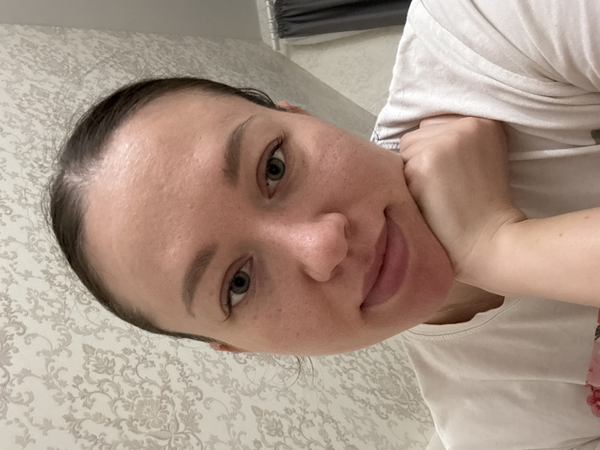 A close-up, slightly rotated selfie of a Caucasian woman with fair skin. She has dark hair pulled back, revealing her forehead. Her eyes are a mix of green and grey. Her left hand rests on her right cheek. She is wearing a light-colored t-shirt. The background consists of a wall with a floral pattern and a window with grey blinds. The location is consistent with an interior space in Ферма, Russia.