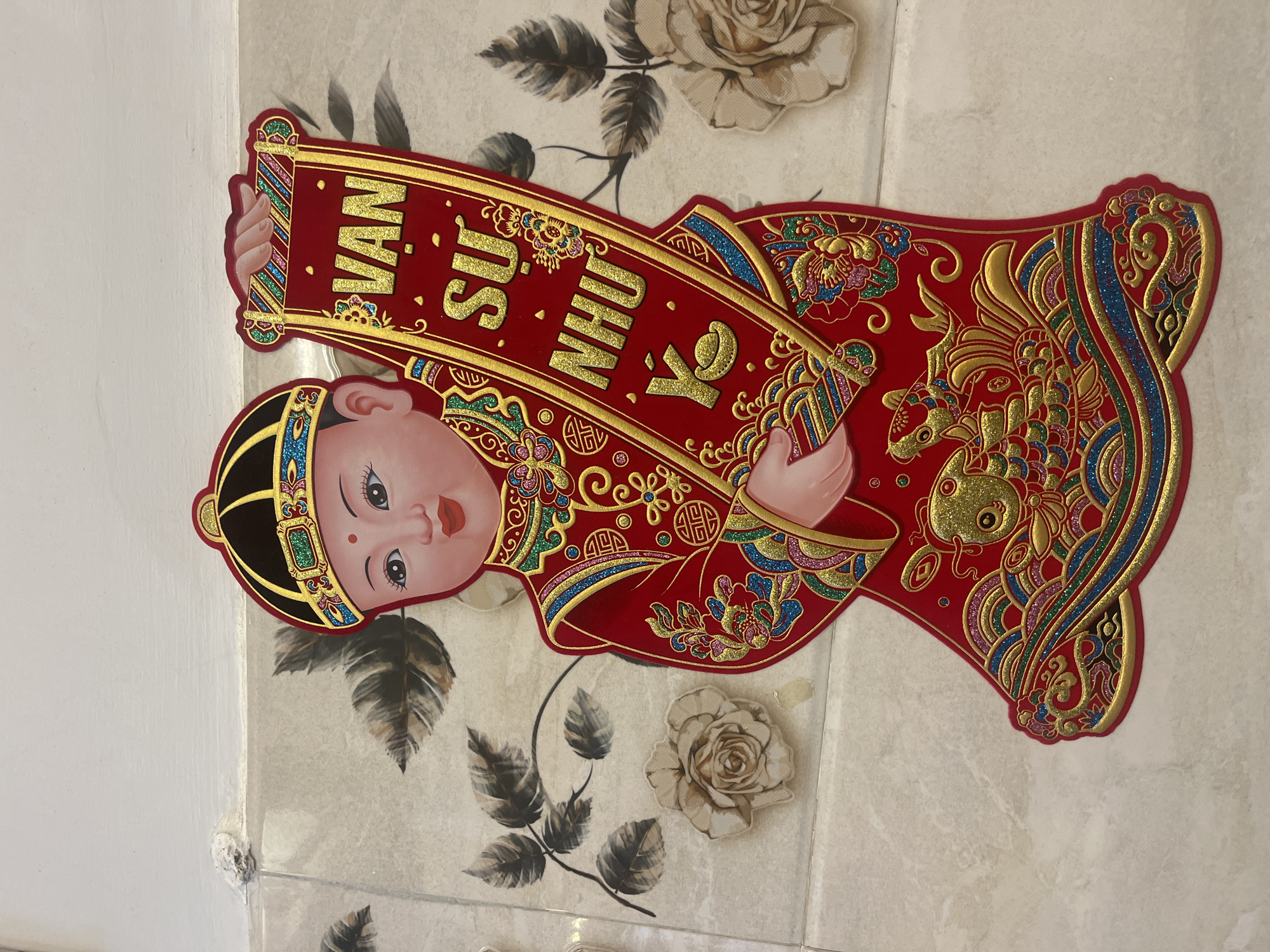 The image shows a decorative cutout of a young boy wearing a traditional red and gold outfit, including a patterned hat. He is holding a red scroll with gold lettering that reads "VAN SU NHU Ý". The cutout is affixed to a wall covered in light-colored tiles with a floral pattern. The scene appears to be indoors, possibly in a residential or commercial setting. The lighting suggests it is daytime. The overall impression is of a festive or celebratory decoration. There is no discernible location within Nha Trang Ward, Vietnam, that can be identified from the image alone.