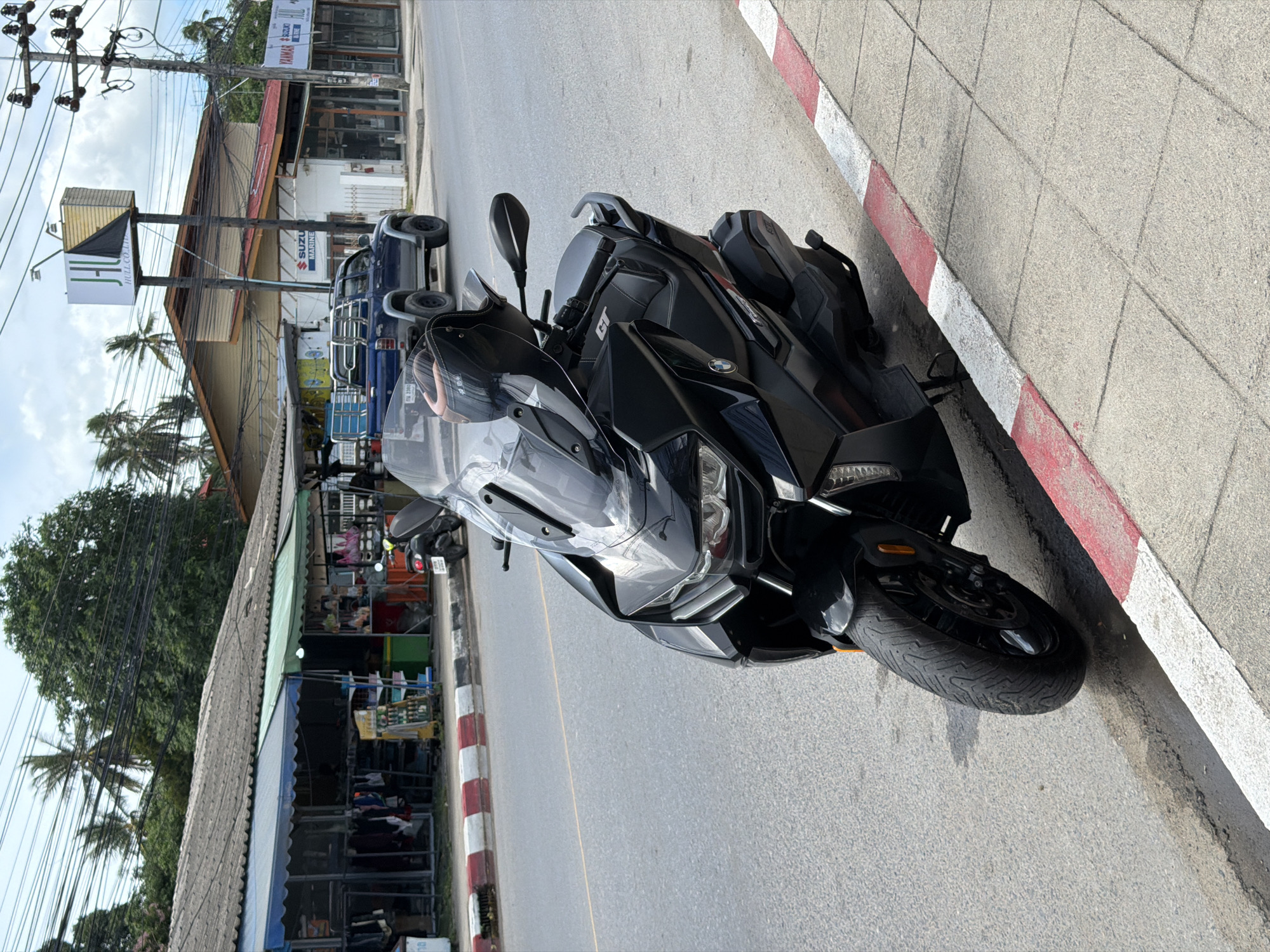 This media file depicts a black motorcycle parked on the side of a street in Baan Mae Nam, Thailand. The motorcycle is the primary subject, angled towards the viewer with its front wheel visible. It appears to be a touring or sport-touring model, characterized by its large fairing, windscreen, and comfortable seating.
The scene is a roadside with a sidewalk marked by red and white painted curbs. Behind the motorcycle, a blue pickup truck is parked. Further in the background, there are several buildings lining the street, suggesting a commercial or residential area. One building appears to be a shop with awnings and merchandise displayed. Overhead, a complex network of power lines and utility poles is prominent, a common sight in many parts of Thailand. Palm trees are visible among the buildings, indicating a tropical or subtropical climate.
The time of day appears to be daytime, with bright sunlight casting shadows. The weather seems to be clear, with some clouds in the sky. The overall atmosphere is that of a typical street scene in a town or village, likely with local businesses and residences.
There is visible text on a sign mounted on a pole in the background, which reads "JH BULL CO.,LTD". Other signs are present on the buildings, but they are less legible in the provided image. The image does not convey any specific emotions or a particular event taking place, other than the general presence of parked vehicles on a street.