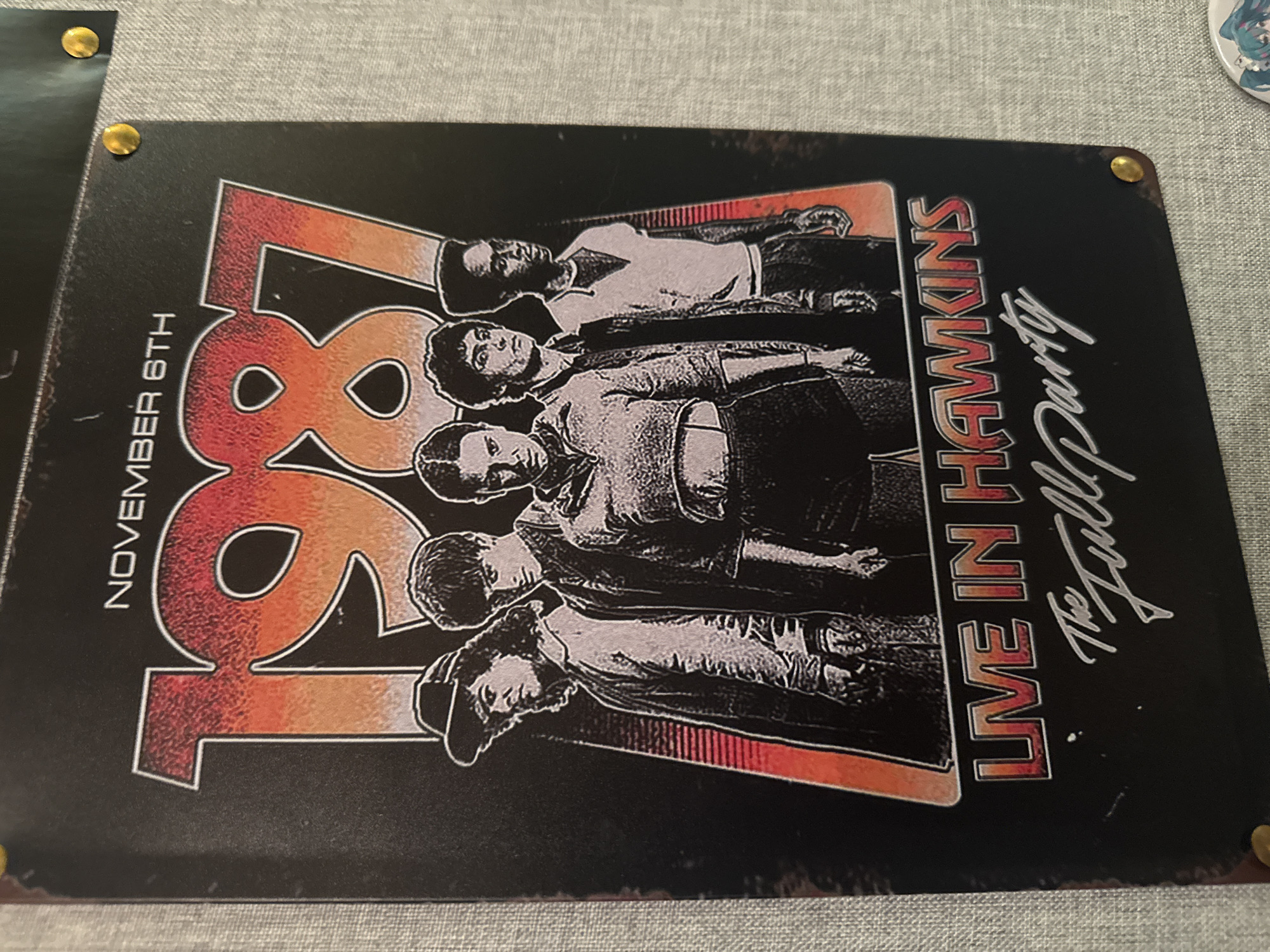 This is a close-up, eye-level shot of a black metal poster pinned to a textured grey wall. The poster features a graphic depicting a group of five young people in what appears to be an 80s aesthetic, with the year "1987" prominently displayed above them in large, stylized lettering. The text "NOVEMBER 6TH" is above "1987," and below the figures, the words "LIVE IN HAWKINS" are in a similar font, followed by "The Full Party" in a script font. The overall impression is that of a promotional poster for an event or a piece of fan art related to the show "Stranger Things," which is set in the fictional town of Hawkins. The lighting is even, and there are no discernible Odesa landmarks or elements of the city visible in the image.