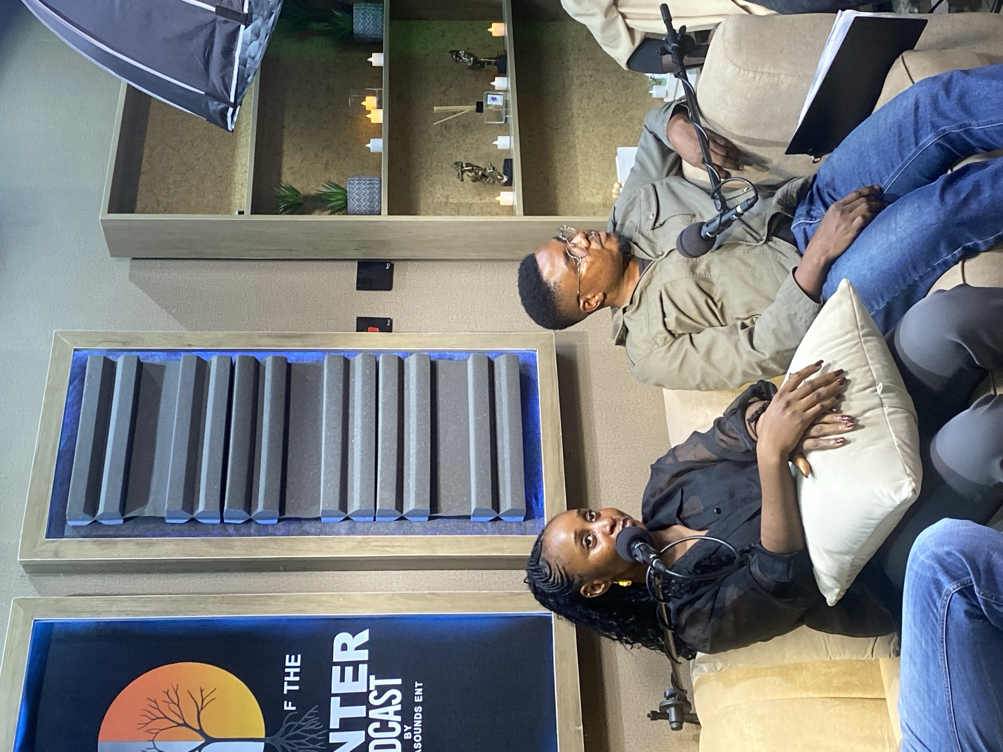 Two individuals, a woman and a man, are seated on a couch in what appears to be a podcast studio. The woman, on the left, has braided hair and is wearing a black sheer top. She is holding a microphone and looking forward. The man, on the right, is wearing glasses and a green jacket. He is looking to his right and has a microphone in front of him. Behind them, on the wall, is a framed poster with graphics and text, as well as acoustic foam panels. To the right, there is a bookshelf with decorative items, plants, and candles. The overall environment suggests a professional recording setup. There is no discernible information within the image that would indicate a specific location in Bar Jirgi Summa, Nigeria.