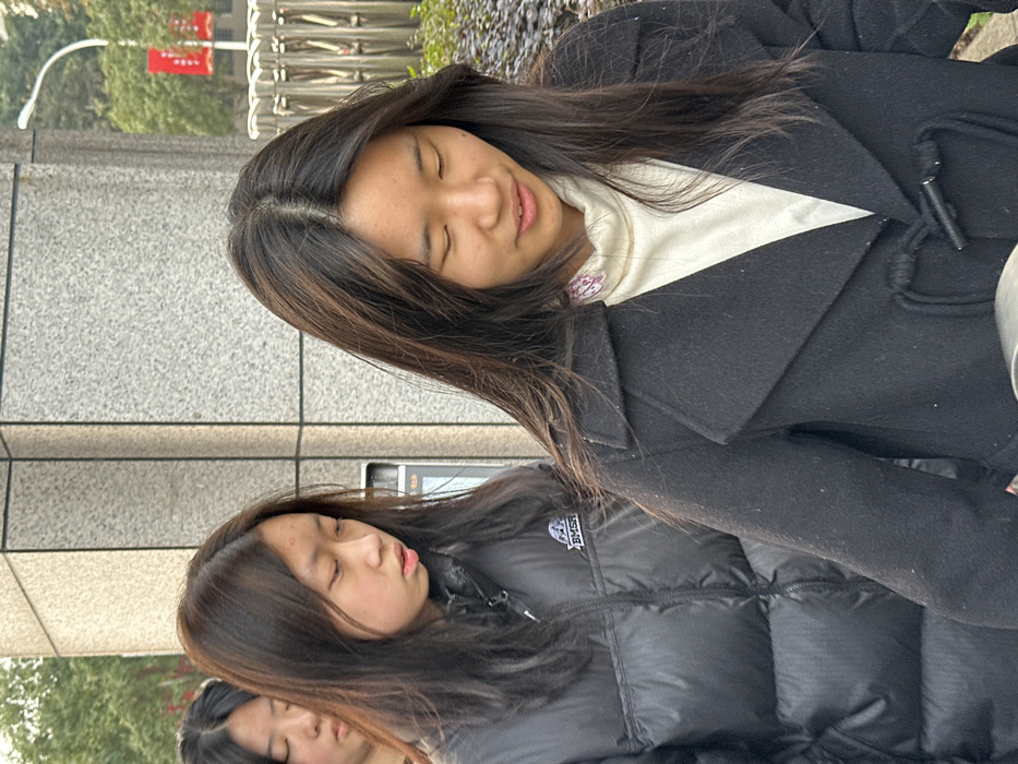 The image depicts three East Asian females walking in succession outdoors in Jiangjin, China. The foremost individual, positioned on the right, wears a black peacoat with two visible toggle closures over a cream-colored turtleneck featuring a small pink and purple floral embellishment on the left collar area. She has long, dark hair parted centrally and her head is angled downwards, with her eyes mostly closed. The second individual, situated directly behind the first and in the center-left of the frame, wears a black puffer jacket with a white 'BMSR' logo and mountain graphic on her left chest. She also has long, dark hair and her head is tilted downwards, with her eyes looking towards the ground. The third individual is partially visible on the far left, with dark hair and a similarly downward gaze. The background consists of a light gray tiled building facade on the left, and a metallic barrier or railing, green foliage, and a white street light pole on the right. The scene is illuminated by soft, ambient daylight.