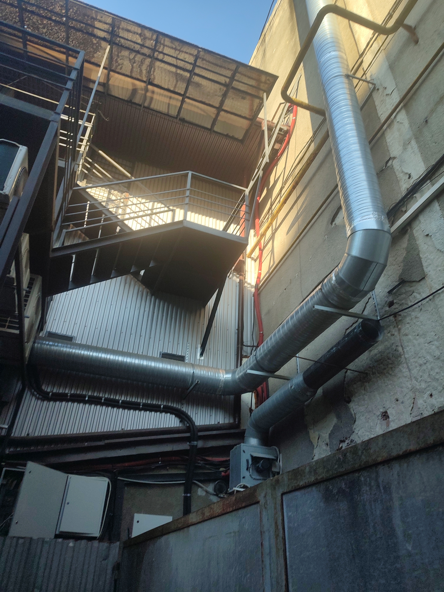 This is a low-angle, wide shot of the exterior of a building with various industrial elements visible. In the foreground, a large, corrugated metal structure is partially visible, topped with a rusty metal beam. Above this, a complex network of metallic pipes runs along the exterior wall of a light-colored, textured building. The pipes vary in size and material, with some being silver and corrugated, while others are dark and smooth. One large silver pipe bends sharply as it descends, supported by a metal bracket. Higher up, a black pipe runs parallel to a silver one.
To the left, a black metal staircase with metal grating and railings ascends towards a covered walkway or roof structure. The roof is covered with what appears to be a translucent material supported by metal beams and a corrugated metal underside. Sunlight filters through the translucent roof, creating diagonal shadows on the walkway and staircase. Some orange piping and red electrical conduit are also visible running vertically along the building and along the staircase. On the left side of the staircase, a white air conditioning unit is partially visible. In the lower left, a light gray electrical panel box with its door ajar is mounted on a corrugated metal surface. The sky above is clear and blue.
This scene is located in Chernivtsi, Ukraine.