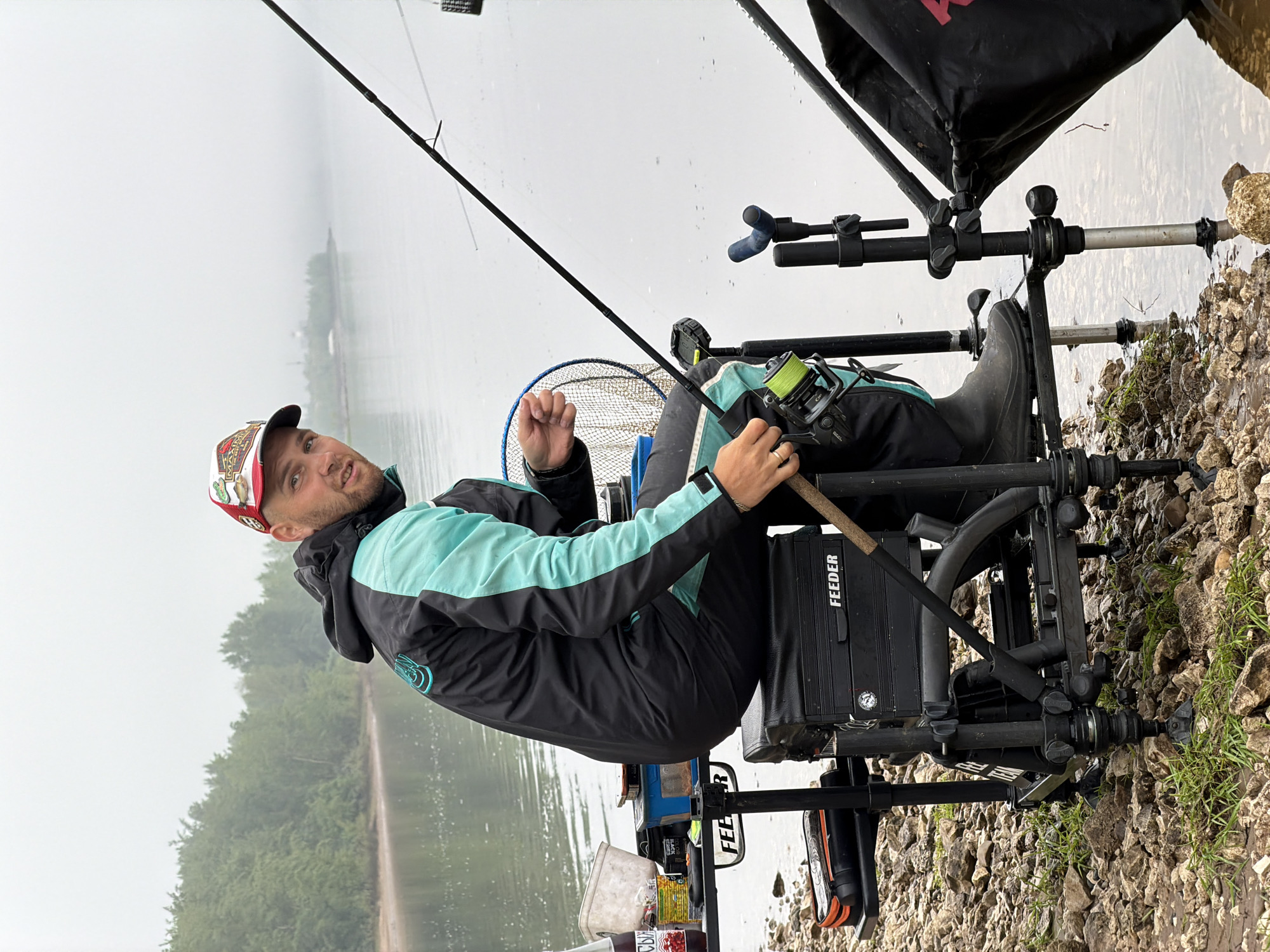 A man is depicted engaged in fishing at a lakeside or riverside location in Noviy, Russia, on what appears to be an overcast day. He is seated on a highly specialized fishing chair or platform, equipped with various attachments for his gear.
The primary subject is the man, who is looking towards the camera with a slight smile or confident expression. He wears a multi-colored cap, a black and light blue (or mint green) outdoor jacket, and dark pants. His right hand is holding a fishing rod, which extends diagonally across the frame, while his left hand is clenched into a fist. A fishing reel with bright yellow line is prominent on the rod. His right foot, clad in a black rubber boot, rests on a component of the fishing station.
The fishing setup is extensive, featuring multiple rod rests, trays, and storage compartments. A black box or container, clearly labeled "FEEDER" in white text, is visible beneath the man's left arm. Another instance of "FEEDER" is seen on a lower box. A blue-rimmed landing net is partially visible behind him. The chair itself is a robust, multi-legged structure, providing stability on the uneven ground.
The setting is a tranquil body of water, likely a lake or river, stretching into the background where a line of distant trees is partially obscured by mist or haze, indicating overcast weather conditions. The shoreline directly beneath and around the fishing station is rocky and pebbly, with some sparse green grass growing among the stones. The water surface appears calm and still, reflecting the muted sky.
The activity is clearly fishing, likely specialized feeder fishing given the specific equipment, with the man poised and attentive to his rod. The overall mood is one of focused outdoor recreation in a natural, somewhat muted landscape.