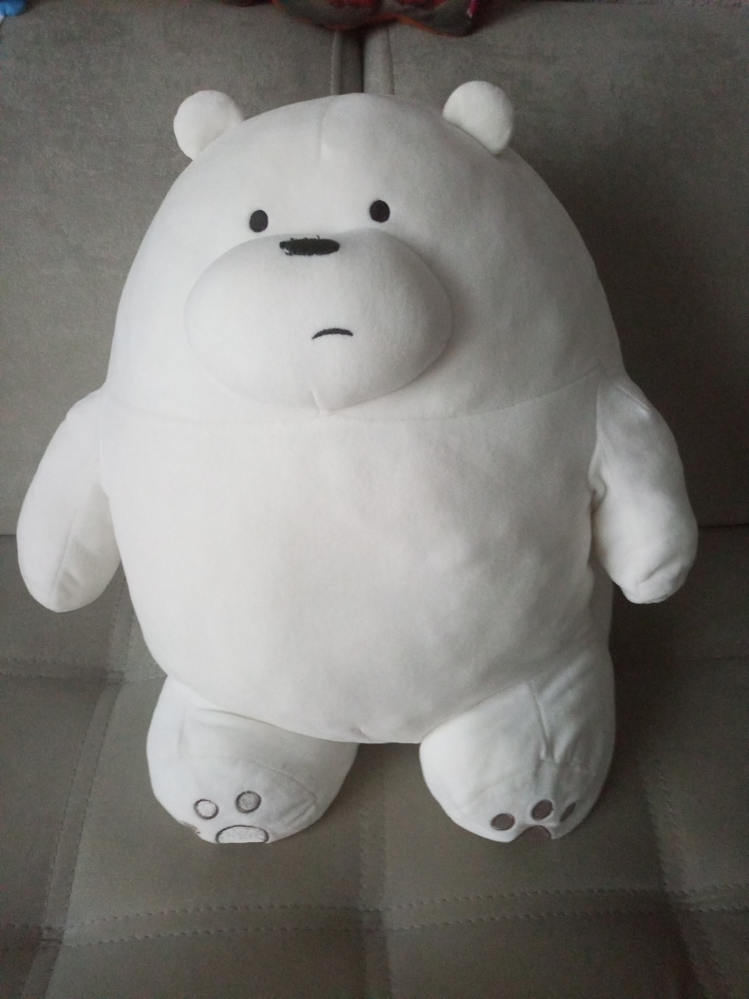 A white plush toy, resembling a polar bear with a black nose and a simple drawn mouth and eyes, sits upright on a textured grey couch. The toy is centrally positioned in the frame and is illuminated by natural light, casting soft shadows. The background consists of the couch and a hint of a patterned fabric in the upper portion of the image. There is no indication of the city of Homyel, Belarus, or any specific location details within the image.