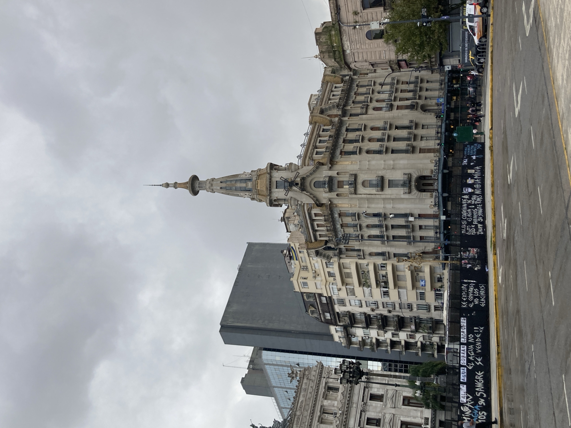 The image captures an urban street scene on an overcast day. A prominent, ornate building with a distinctive spire and intricate architectural details dominates the left side of the frame. Its facade is a light, somewhat weathered stone color, featuring numerous windows and balconies. To the right of this building, a modern, dark grey building with reflective windows is partially visible, contrasting with the older architecture.
In the mid-ground, a wide street runs vertically, featuring road markings and a yellow dividing line. Parallel to the street, a dark wall or barrier is heavily covered in white graffiti. A few people are visible on the sidewalk behind this wall, along with a white van and a smaller dark vehicle parked. Streetlights and some greenery are also present. The sky is uniformly grey and cloudy, indicating an overcast day.
Visible text provides significant clues. On the ornate building, "CAFETERIA DEL MOLINO" is legible, referring to the famous Confitería del Molino in Buenos Aires, Argentina. A street sign states "SABADO 5 DE SEPTIEMBRE." The graffiti on the wall conveys strong social and political messages in Spanish, including: "FALTA AGUA SOBRAN LADRONES" (Lack of water, too many thieves), "EL AGUA NO SE VENDE" (Water is not for sale), "AGUA SIN SANGRE" (Water without blood), "KE EXPLOTE EL CONGRESO NO LOS GLACIARES!" (Let Congress explode, not the glaciers!), and mentions of "MILESI CONTAMINANKS" (referring to politician Javier Milei and contamination) and "TRUMP SU SANGRE TODYS NO LA MINA" (possibly anti-mining and anti-Trump sentiment). These texts suggest local protests or expressions of discontent.