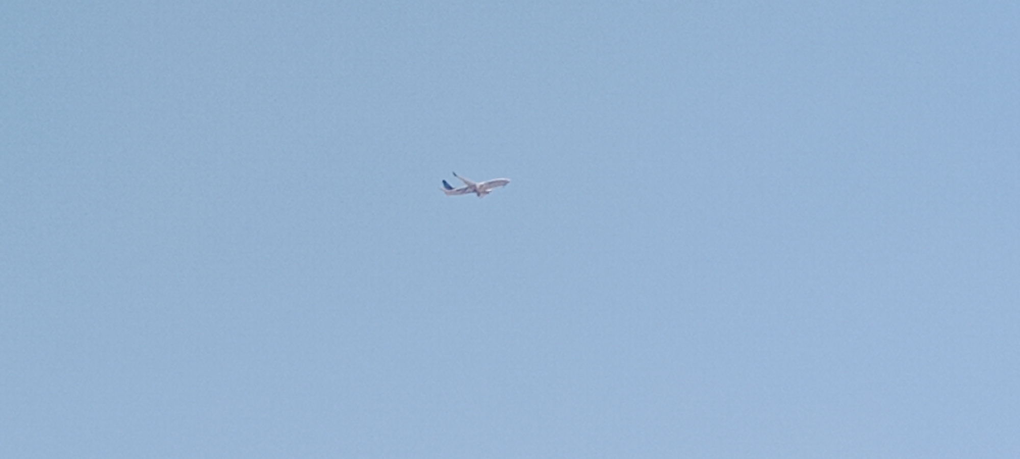 A single airplane is visible against a clear blue sky. The airplane is white with blue accents on its wings. It is in flight and appears to be ascending. The image does not provide enough context to determine the location within Colmenar Viejo, Spain.