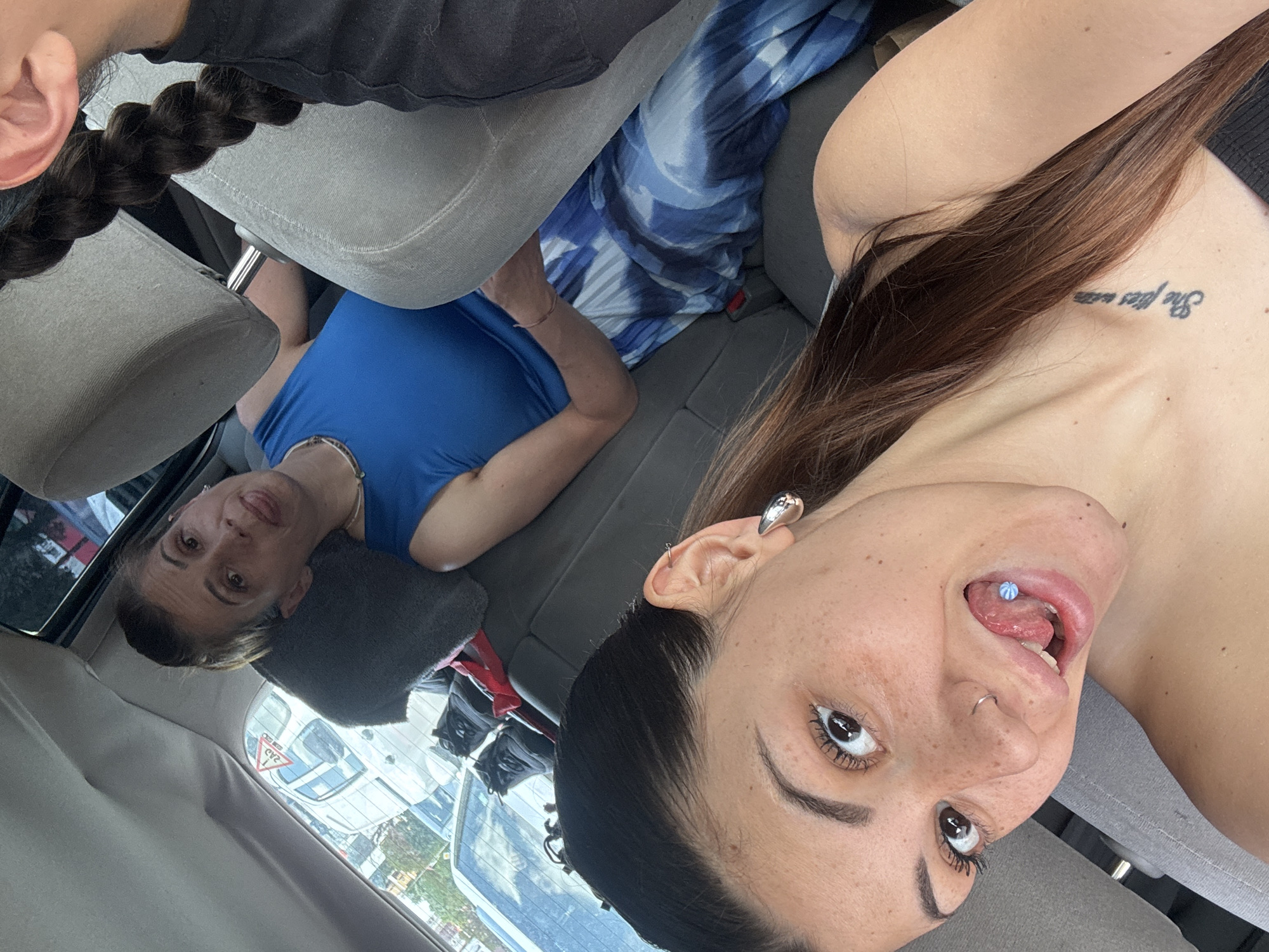 The image depicts the interior of a vehicle, likely a car, with three individuals present.
In the foreground, a young adult female, possibly the front passenger, takes a selfie. She has fair skin with visible freckles, dark brown hair, and dark eyebrows with mascaraed eyelashes. She displays a silver nose ring on her left nostril, a silver teardrop earring in her left ear, and an upper ear piercing in her right ear. She wears a dark, possibly black, strapless top, and a tattoo reading "She flies with..." is visible on her right collarbone. She is looking directly at the camera with her tongue extended, revealing a light blue beaded tongue piercing.
In the midground, seated in the rear passenger seat directly behind the foreground subject, is an adult female. She has fair skin and her hair is pulled back. She wears a bright blue sleeveless top and a blue and white tie-dye patterned skirt or trousers. A thin necklace is visible around her neck. She is also looking towards the camera with her tongue extended. A grey cushion is positioned behind her head against the car's headrest.
Partially visible on the far right of the image, beyond the front passenger's headrest, is a third individual, likely the driver. Only a dark, tightly braided ponytail and a portion of an ear, wearing a dark top, are discernible.
The car's interior features grey fabric seats and headrests, along with dark grey plastic trim. A seatbelt buckle is visible. On the rear parcel shelf, behind the rear passenger, black footwear and a bag are present.
Through the vehicle's rear window, an urban street or parking lot is visible with other cars and trees. A white car in the background displays a red and white triangular sign with the letters "ZAD" visible through its rear window. This type of sign is indicative of Bulgaria. Given this detail, the scene is located in Bulgaria, specifically suggested to be in the city of Sofia.