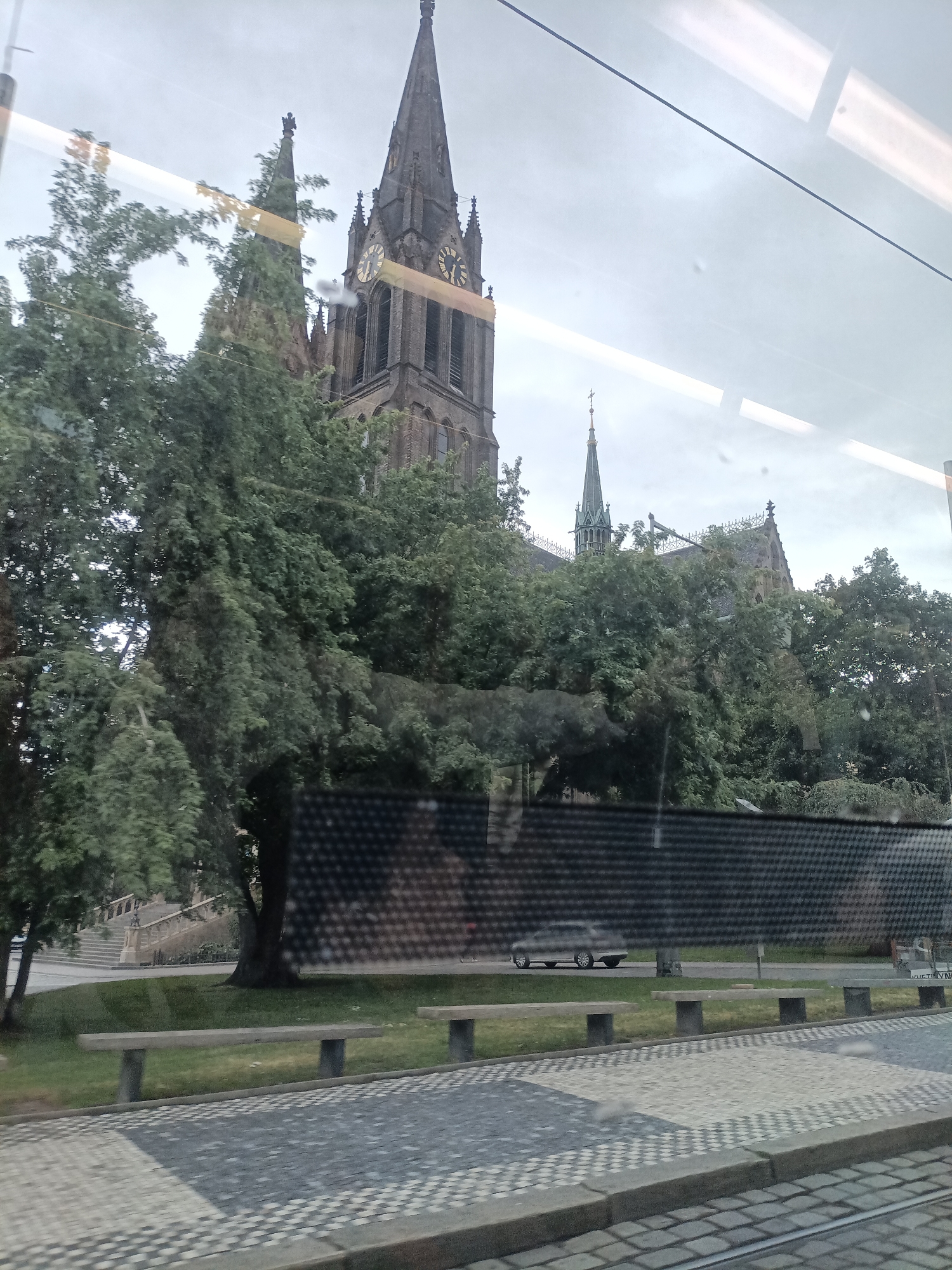 The image shows a view through a window, likely from a moving vehicle, of the St. Vitus Cathedral in Prague, Czechia. The cathedral's gothic spires and clock tower are visible above a dense canopy of green trees. In the foreground, there are paved walkways and benches. A silver car is visible in the middle ground, driving on a road behind a dark, textured fence. The sky is overcast, suggesting a cloudy day. The reflection of lights and lines from the window are also present in the image.