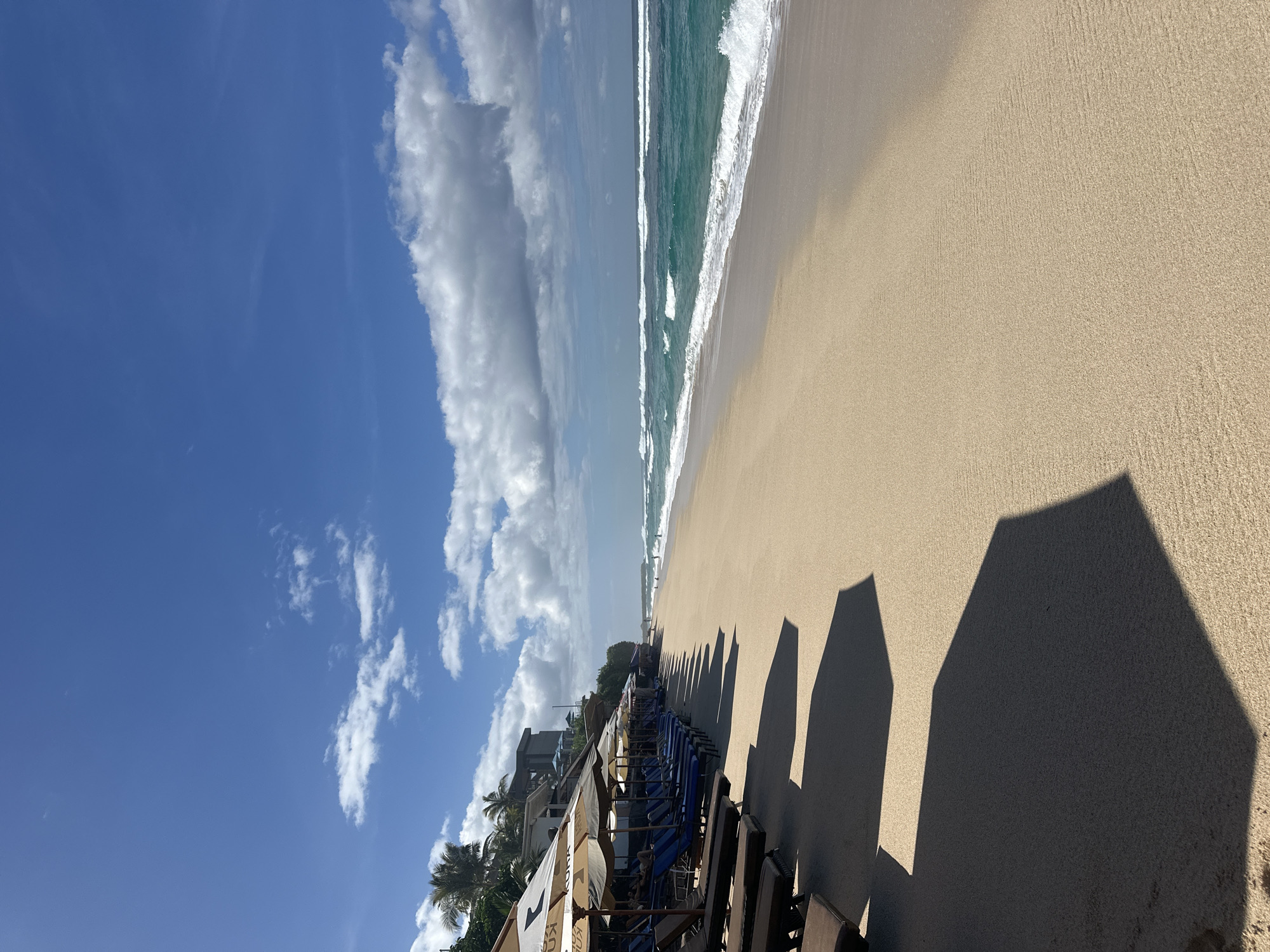 The image shows a sunny beach in Kuta Selatan, Indonesia. The sky is blue with scattered cumulus clouds. The ocean has turquoise water with white waves breaking on the shore. The beach is sandy, and there are several rows of beach chairs with umbrellas casting long shadows on the sand. Some palm trees are visible near a building in the background.