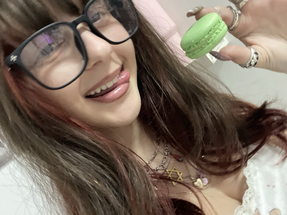 A woman with red-streaked brown hair smiles, her tongue protruding slightly. She wears large, black-framed glasses. A green macaron is held in her right hand adorned with multiple rings. She wears a silver chain necklace with a Star of David pendant and other charms. A white, lace-trimmed garment is visible at the bottom of the frame. The background is a soft pink.