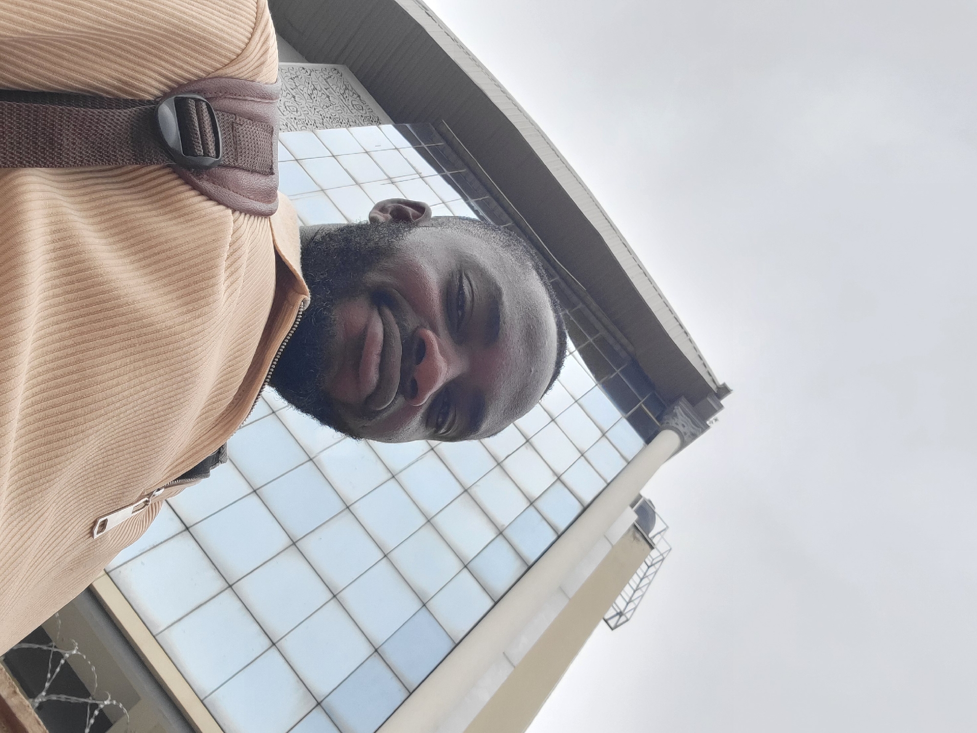 An outdoor, low-angle portrait captures a man with dark skin and a short beard, looking directly at the camera with a subtle, content smile. He is wearing a light tan or beige corduroy jacket with a silver zipper pull visible on the left side of his chest. A brown, textured backpack strap with a black plastic buckle crosses his upper left shoulder.
The photograph is taken from a perspective looking upwards, positioning the man in the foreground while a modern-looking building dominates the background. The building features large, gridded glass windows that reflect a bright, overcast sky. Architectural details are visible on the building's facade, including a curving concrete or stone structure and a section with an ornate, white patterned design. The sky is uniformly light grey, indicating an overcast day. In the lower left corner of the frame, strands of barbed wire are visible, suggesting a security fence or barrier. The overall lighting is soft and diffused. The image is oriented sideways, appearing rotated 90 degrees counter-clockwise, as if the original shot was taken with the camera in landscape mode while looking upwards. The setting is an urban environment in Jos, Nigeria. No text is visible in the image.