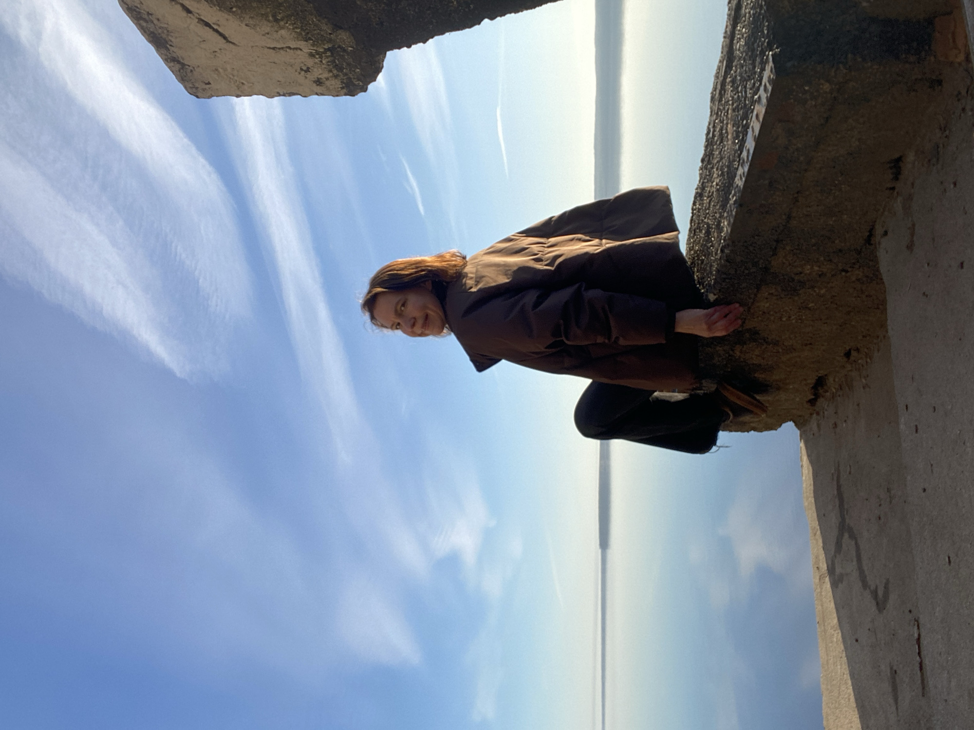 The image, originally rotated 90 degrees clockwise, presents a woman posing on a concrete structure, overlooking a vast body of water under a bright blue sky.
The central subject is a woman with brown hair, wearing a dark brown, possibly padded, jacket and dark trousers. She is positioned slightly to the left of the frame's center, leaning forward with her body angled, and her left hand gently resting on the concrete wall beside her. She is looking directly at the camera with a subtle, friendly smile, conveying a sense of ease and contentment.
The setting is outdoors during the daytime, under a clear and sunny sky. The sky is a vibrant blue, featuring delicate, streaky white clouds that stretch across the expanse. Below the horizon, a large body of water, likely a river or reservoir given the Chaikovsky, Russia context, extends into the distance, meeting the sky in a hazy line. The foreground and right side of the image are dominated by rough, textured concrete structures, suggesting a man-made environment such as a pier, dam, or industrial ruin adjacent to the water. These structures show signs of weathering.
No legible text or identifiable markings are visible on the concrete or in the background. The scene captures a tranquil moment, with the woman as the focal point against the expansive natural backdrop of sky and water.