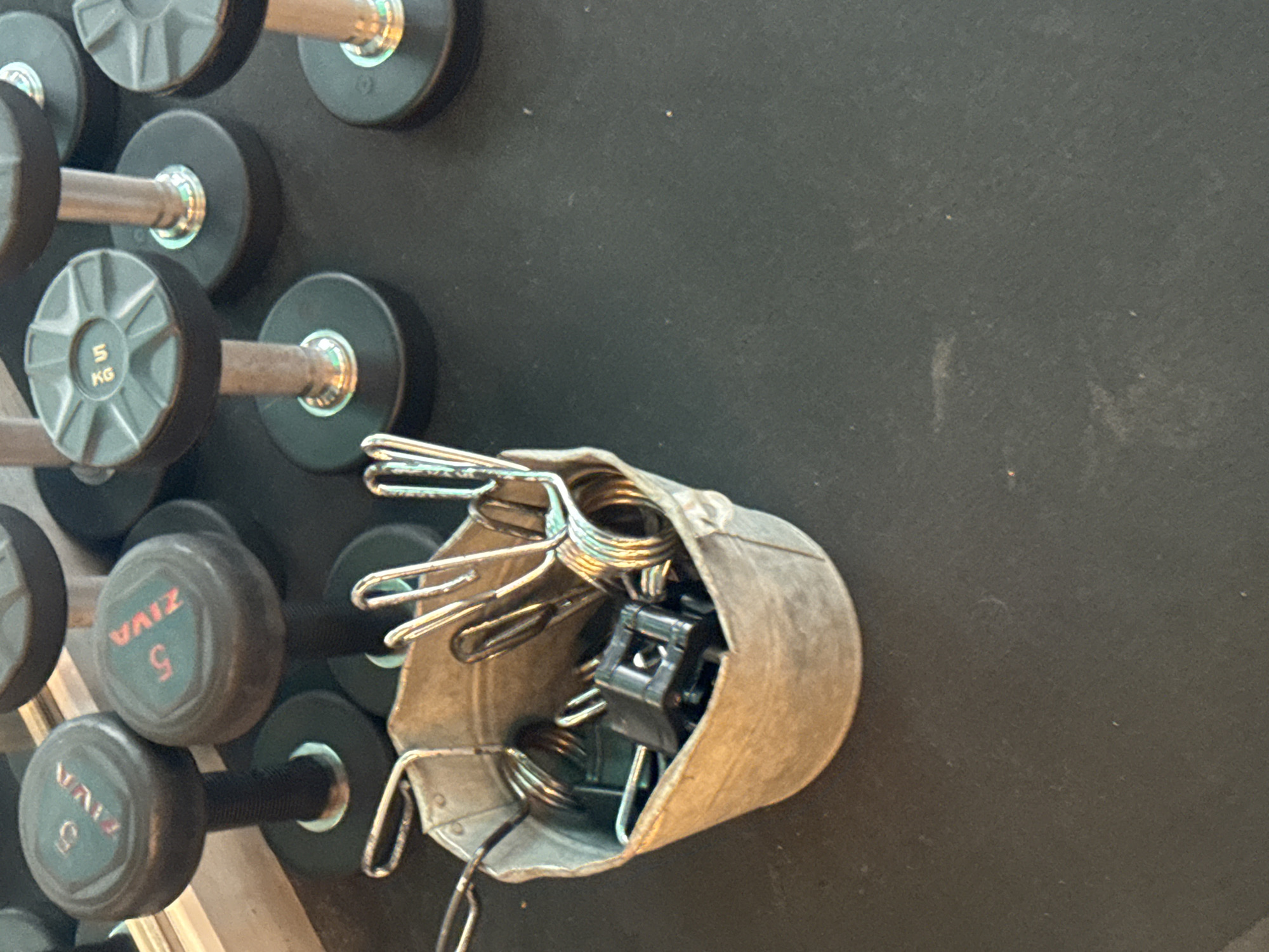 This image depicts a collection of weightlifting equipment, specifically dumbbells, arranged on a dark-colored floor, likely in a gym. In the foreground, a distressed metal bucket holds several spring-loaded dumbbell collars. The background features multiple dumbbells of varying weights. Two dumbbells on the left side of the frame have "ZIVA 5" inscribed on their ends, indicating they are 5-kilogram weights. Other dumbbells visible have similar inscriptions, including "5 KG". The overall environment suggests a fitness facility. There is no indication of the city of Murcia, Spain.
